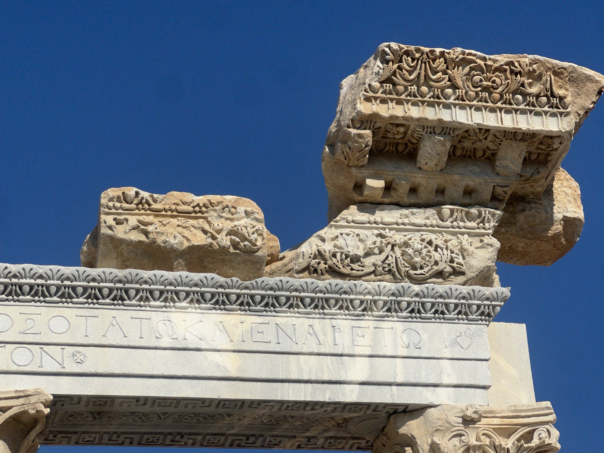 A close-up, eye-level, medium shot captures fragmented ancient ruins against a clear, deep blue sky. The ruins are made of weathered, light-colored stone and feature intricate carvings. In the foreground, a stone archway is visible, decorated with a repeating geometric pattern and what appears to be Greek lettering. Above the archway, two separate stone blocks rest, also adorned with elaborate floral and scrollwork carvings. The larger block on the right juts out prominently, showcasing detailed relief work. The overall impression is one of a sun-drenched archaeological site, with the vibrant blue sky emphasizing the texture and age of the stone structures.