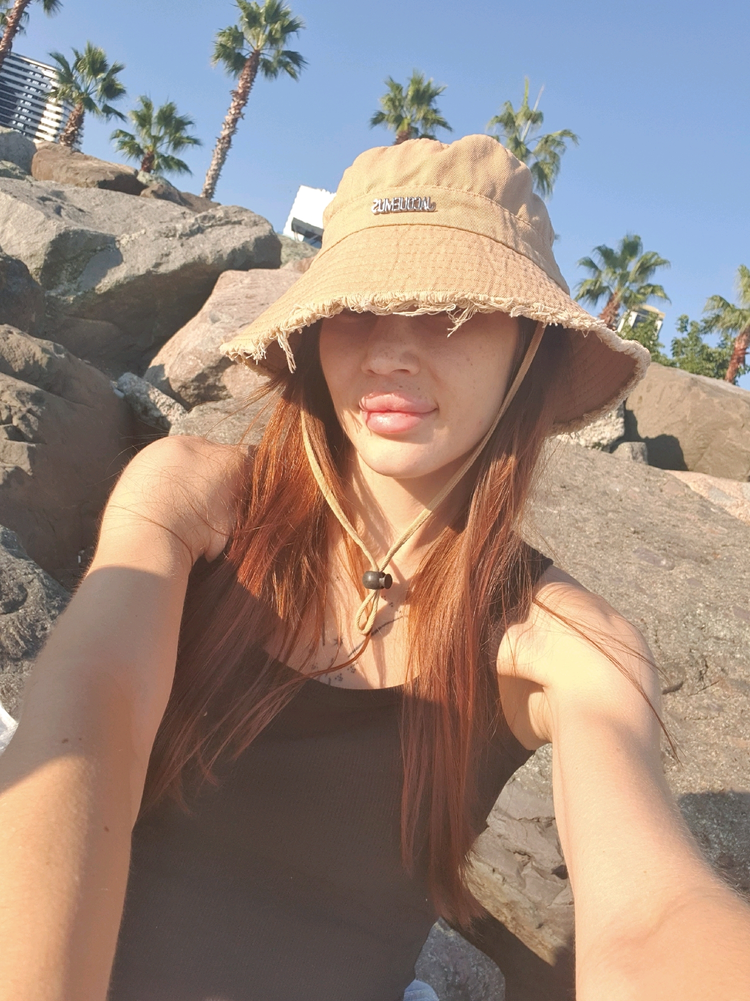 The image is a selfie taken outdoors, with the subject, a woman with long auburn hair, wearing a tan bucket hat and a dark tank top. She is sitting on large rocks, with palm trees and buildings visible in the background. The lighting suggests it is daytime, possibly late afternoon, with strong sunlight casting shadows. The overall scene appears to be in a coastal or rocky area within a city, with elements suggesting a warm climate. The location is identified as Batumi, Georgia.