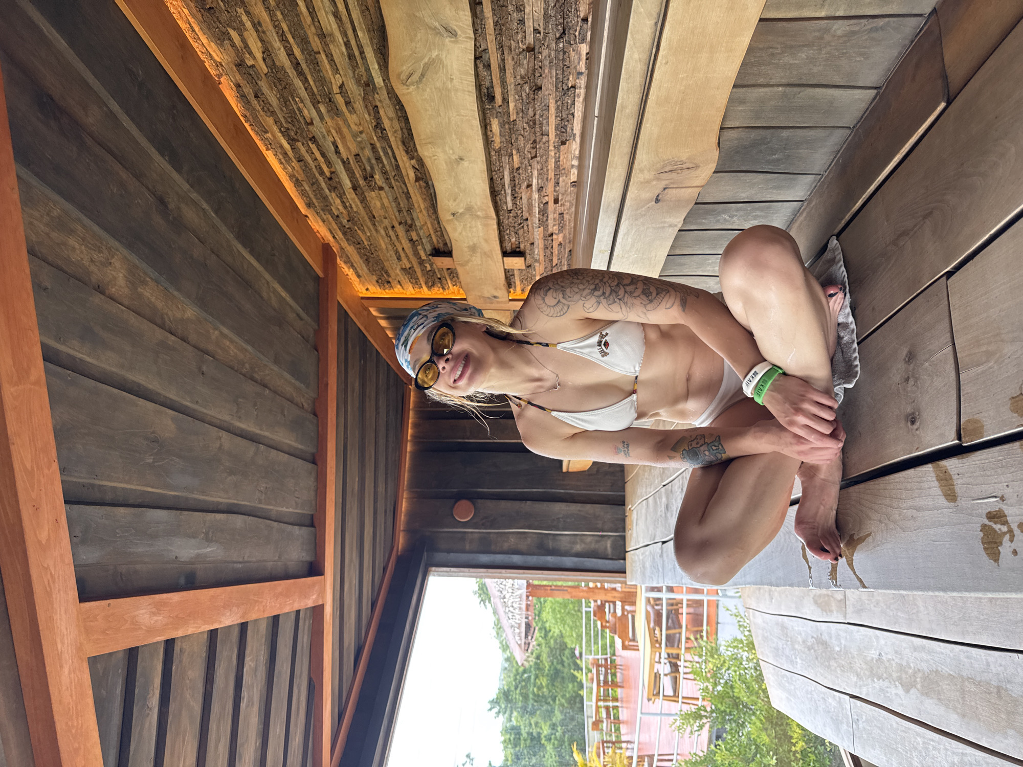 A young woman is seated on a light-colored wooden deck or floor, leaning against a darker wooden wall, within a structure primarily constructed from wood. She is looking directly at the camera with a cheerful smile. She wears a white bikini top, yellow-tinted sunglasses, and a patterned headscarf or bandana. The text "BILLABONG" is visible on her bikini top. Multiple tattoos adorn her arms, and a green wristband with "KAI E" visible on it is on her left wrist.
Her skin and the surrounding wooden surfaces appear wet with water droplets, suggesting she has recently been in water. A small gray towel is placed beneath her. The interior architecture features various wooden textures, including smooth planks, rugged bark-like panels on the ceiling, and reddish-brown support beams. In the background, an opening reveals an outdoor scene with lush green trees and what appears to be resort-style buildings, indicating a natural, tropical environment. The scene is illuminated by natural daylight, conveying a relaxed and pleasant atmosphere in Kathu, Thailand.