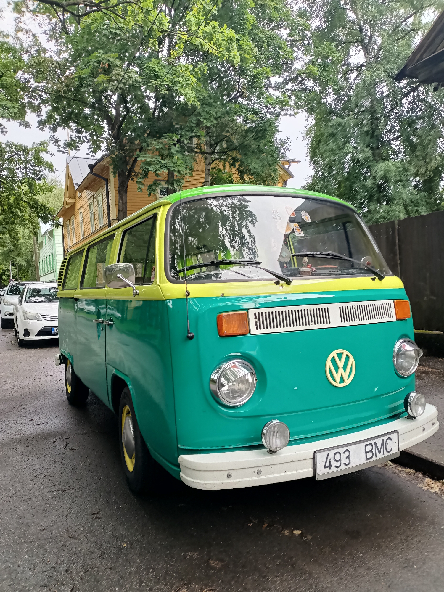 A bright green and yellow Volkswagen Type 2 van is parked on a street in Tallinn, Estonia. The van is the primary subject, positioned in the foreground and angled towards the viewer. It has a classic design with a split windshield, round headlights, and a chrome grille with a yellow Volkswagen logo. The van's license plate reads "493 BMC". Behind the van, a row of white cars is visible, also parked along the street. The street is paved and appears wet, suggesting recent rain. In the background, traditional wooden houses with yellow and light green facades line the street. Lush green trees with dense foliage are abundant, creating a canopy over the street and houses. The sky is overcast. The scene depicts a residential street in Tallinn, characterized by its historical architecture and abundant greenery.
