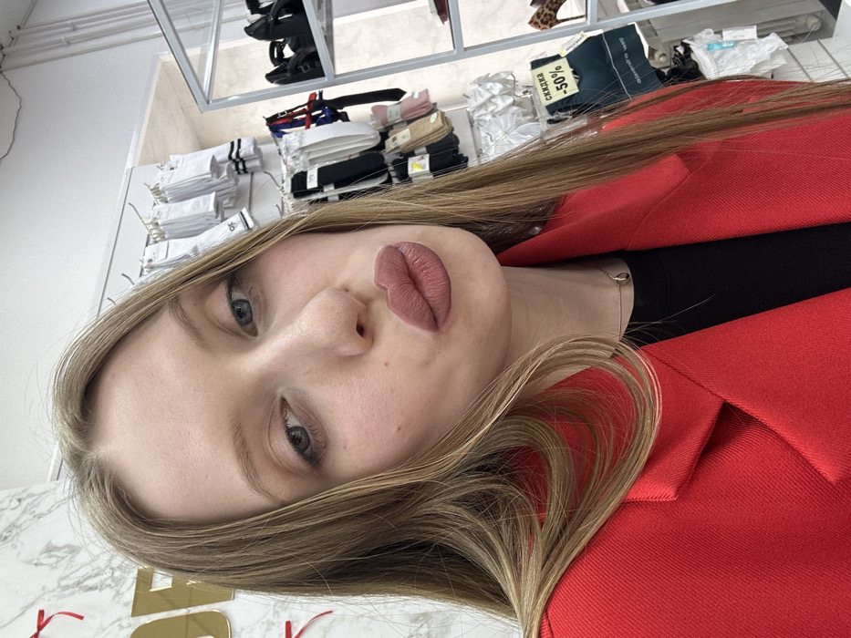 A young female subject is depicted in a selfie, positioned within a retail clothing and accessories store. She has long, straight light brown hair, a fair complexion, and blue eyes, and is wearing a bright red blazer over a black top with a delicate necklace. Her lips are pursed in a "duck face" expression, coated with matte rosy-brown lipstick, and she looks directly at the camera.
The background shows a store interior with white walls. To the left, a wall featuring a subtle grey marble-like pattern displays gold-colored dimensional lettering, partially visible as "DE" or "OF," accompanied by red decorative accents. To the right, various socks and hosiery in white, black, and nude tones are displayed on hooks. A white sign with red text reading "СКИДКА -50%" (DISCOUNT -50%) is visible. A metallic shelving unit on the far right holds dark shoes, including a black pump and a leopard-print high heel. The ceiling is white with minor visible cracking. The specific location within the city of Chaikovsky, Russia, is not discernible from the image, beyond confirming a Russian-speaking environment due to the signage.