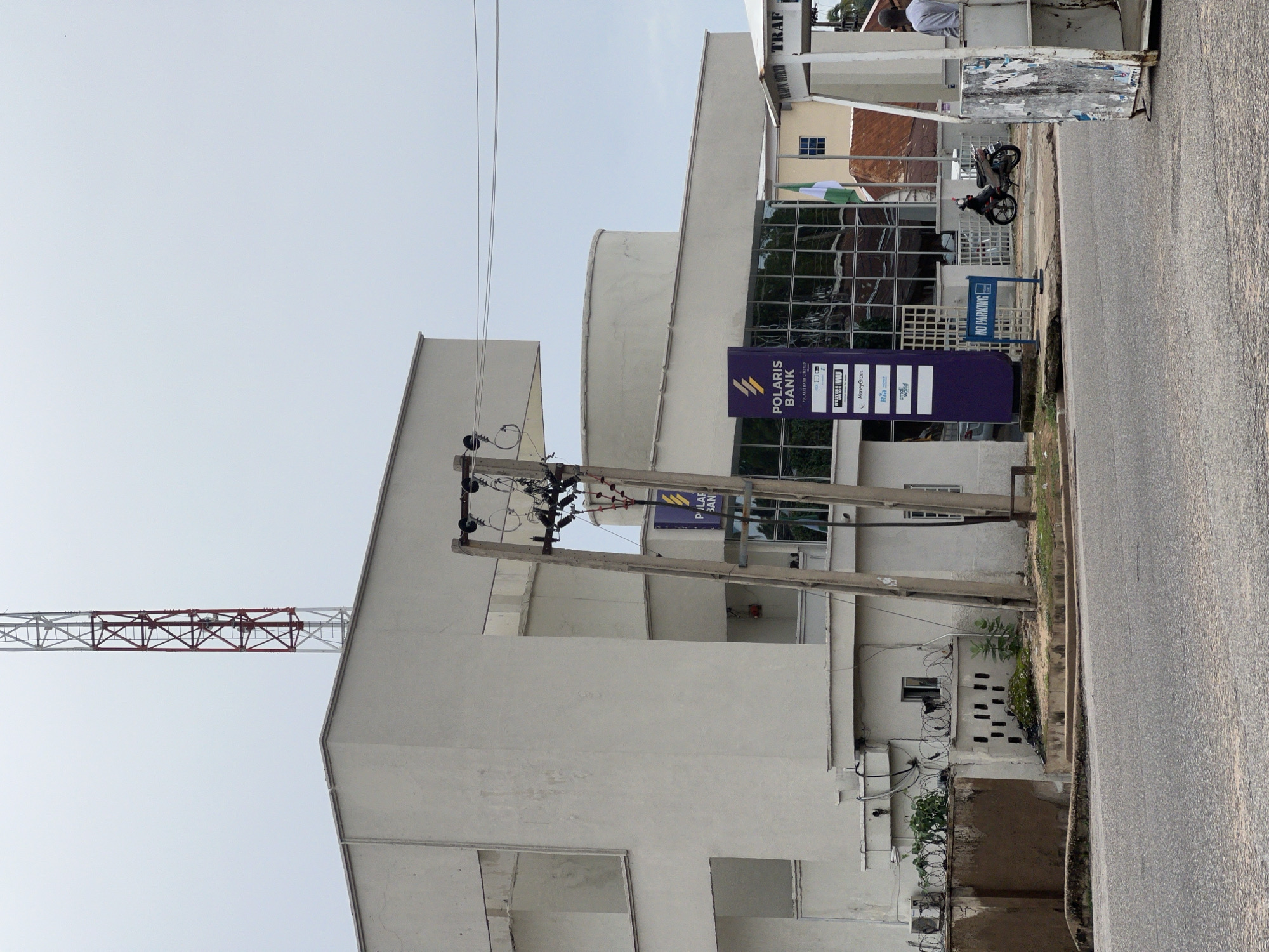 The image shows a street scene in Jalingo, Nigeria, with a focus on a modern building housing a Polaris Bank branch. A tall, red and white telecommunications tower stands in the background against an overcast sky. The bank building has a distinctive geometric design with a curved section. In front of the bank, a purple sign displays the Polaris Bank logo and names of associated services like Western Union and MoneyGram. A "NO PARKING" sign is also visible. A motorcycle is parked on the sidewalk, and a man in a grey shirt is standing in a small white booth-like structure on the right side of the image. Electrical poles and wires crisscross the scene, and a Nigerian flag is flying near the bank. The foreground features a paved road. The overall impression is that of a typical urban street in Nigeria.