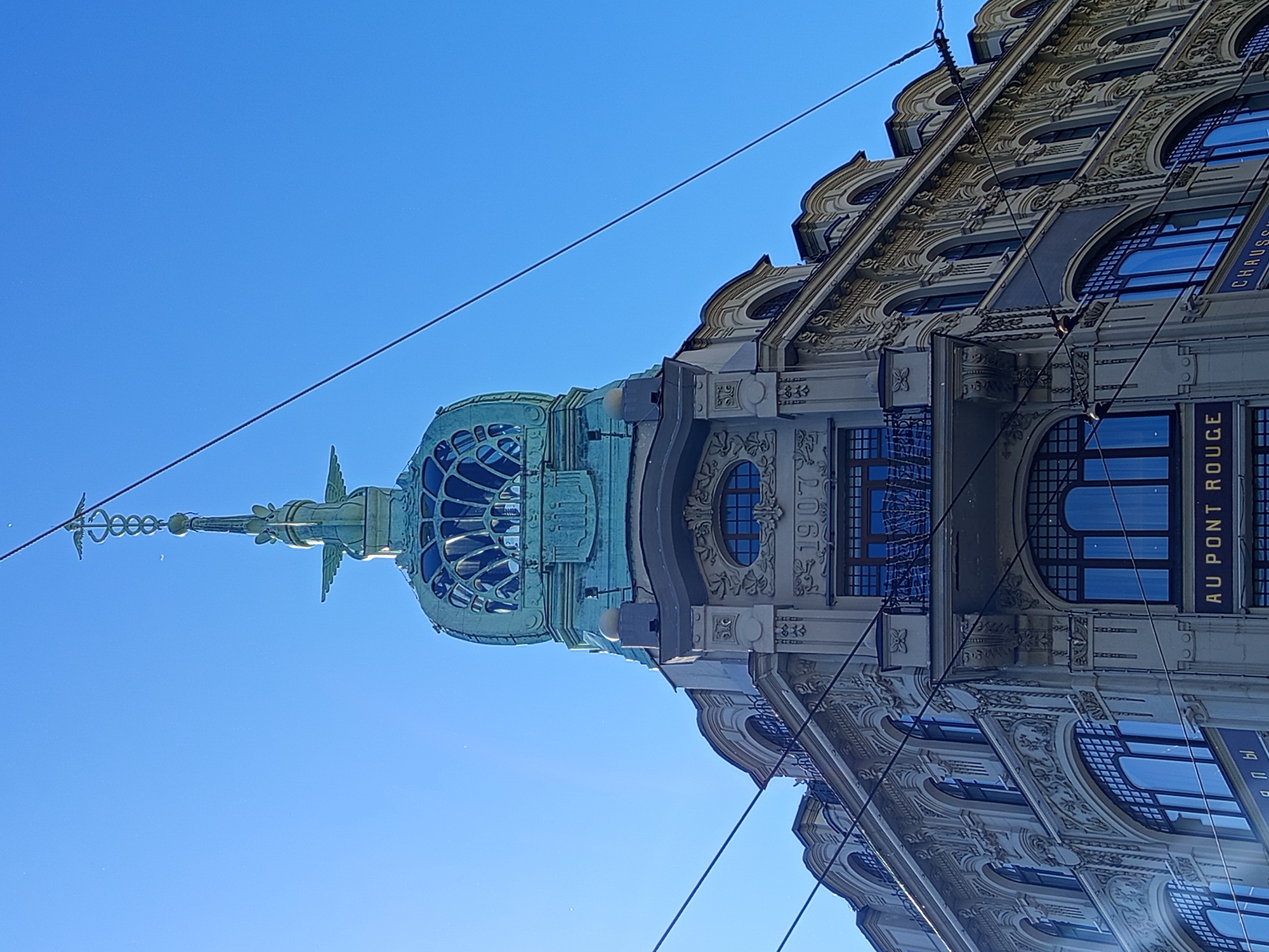 This media file is a photograph of an ornate building with a distinctive copper-colored dome against a clear blue sky. The building exhibits intricate architectural details, including decorative carvings and arched windows. Some windows are reflective, showing the sky and possibly surrounding elements. The perspective is from below, looking upwards towards the dome and the upper levels of the building.
Notable details include the presence of power lines or cables crisscrossing the sky, obscuring parts of the view and suggesting an urban environment. The building's facade is detailed with patterns and moldings, indicative of a historic architectural style. Some text is visible on banners or signs on the building. One sign reads "AU PONT ROUGE" in white letters on a dark background, and another partially visible sign appears to have Cyrillic lettering. The year "1902" is also visible in raised numerals on the building's facade.
The lighting suggests it is daytime, with bright sunlight illuminating the building and creating a sharp contrast against the deep blue sky. The weather appears clear and sunny. The overall impression is one of historical grandeur and architectural beauty within an active urban setting. The image captures a slice of Saint Petersburg's architectural heritage. There are no people visible in the frame, focusing entirely on the structure and its surroundings.