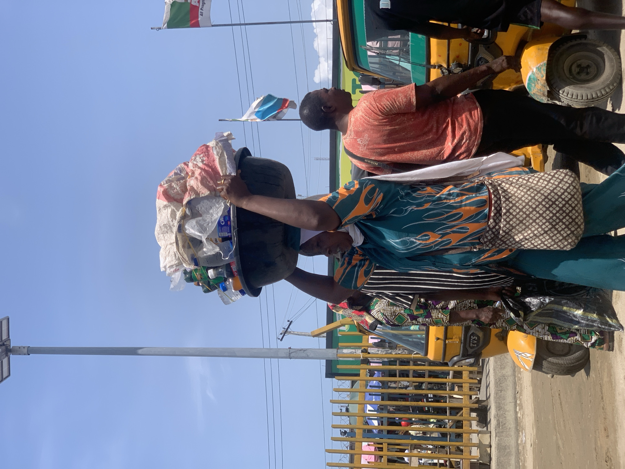 This media file captures a vibrant street scene in Lagos, Nigeria, under a bright blue sky on a clear day. The image is originally oriented sideways but depicts an upright view.
In the foreground, a woman walks while expertly balancing a large, dark basin filled with various plastic bottles, bags, and other goods on her head, indicating she might be a street vendor or hawker. She wears a patterned tunic-style top in teal with orange and black designs, a white headwrap, and a traditional patterned skirt in green, black, and white. She also carries a patterned shoulder bag. Beside her, another person, partially obscured, is also walking, wearing a striped top and a patterned wrapper or skirt.
To their right, a man in a salmon-orange t-shirt and black trousers stands near a yellow motorized tricycle, commonly known as a "keke Napep" in Nigeria. He wears a strap around his neck with the word "Nikon" visible. Another person, mostly out of frame, is visible near the front of the tricycle. The tricycle has "TVS" visible on its side, a common brand for these vehicles.
In the background, two flags are visible on poles against the clear sky. One flag displays the green and white colors of the Nigerian flag, and also features visible text that appears to read "All Progre...", likely referring to a political party (e.g., All Progressives Congress). Another flag, partially visible, has blue, white, and red colors. A utility pole with what appears to be a solar panel or light fixture stands on the left. Faint power lines crisscross the sky. A yellow metal fence or gate runs along the street in the midground, with glimpses of buildings or structures behind it. The ground consists of a paved road or concrete area leading to a dirt shoulder. The overall impression is one of daily life and commerce on a busy African street.