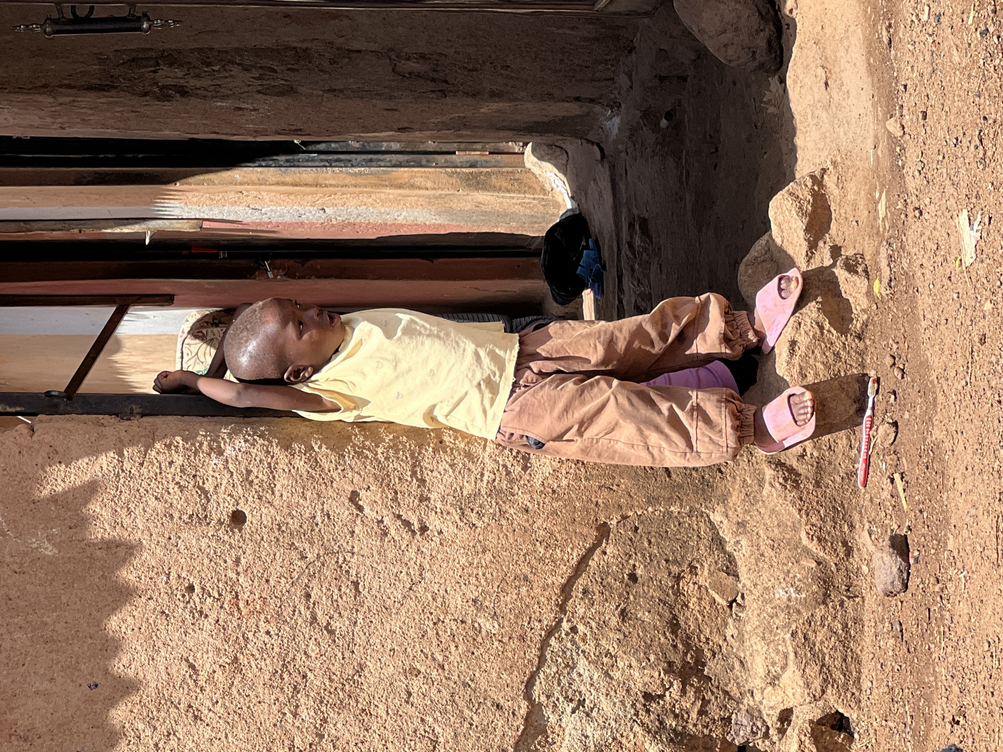 A child is lying on a surface between two walls of a building in Gura Top, Nigeria. The child is wearing a yellow shirt, brown pants, and pink sandals. A toothbrush is lying next to the child's feet.