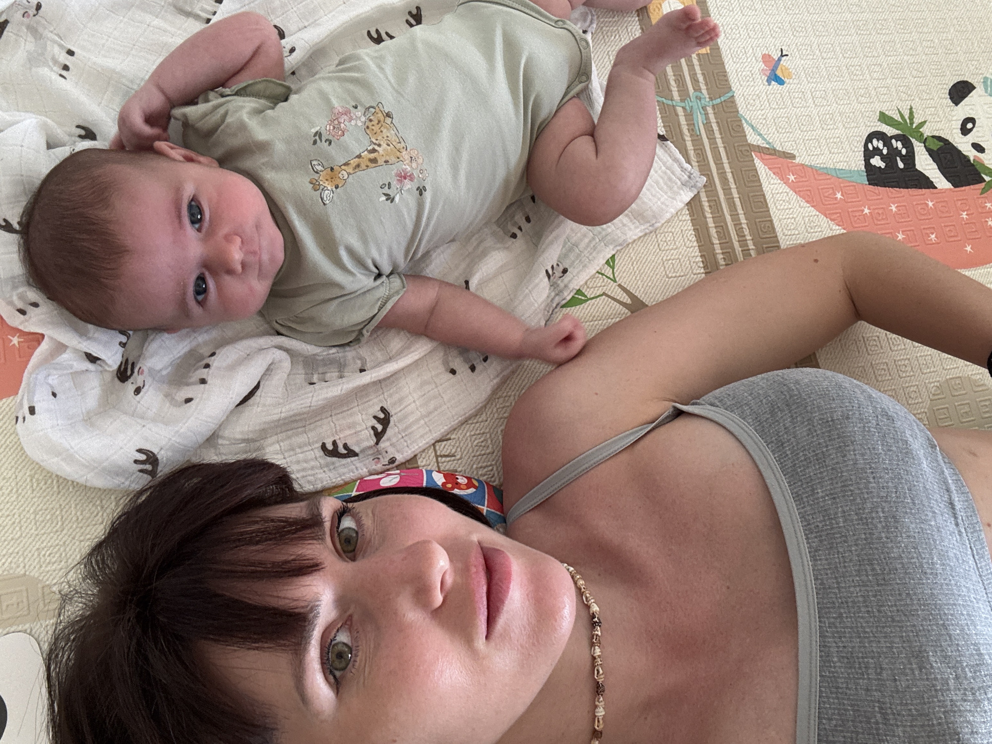 This is a top-down, selfie-style photograph featuring two subjects: a woman and a baby. The woman, positioned in the foreground, is lying on her back and looking up at the camera. She has dark brown hair with bangs and striking green eyes. She is wearing a grey tank top and a beaded necklace. The baby, lying to her left and slightly above, is also on their back, wearing a light green onesie with a giraffe and floral design. The baby has fair skin, dark hair, and blue eyes, and appears to be looking towards the camera as well.
They are both lying on a soft surface. Beneath the baby is a white muslin blanket with a pattern of small, dark animal silhouettes. The woman and baby are on top of a colorful play mat with a textured pattern. The mat features an illustration of a panda eating bamboo in a hammock and a butterfly.
The lighting suggests it is daytime, and the overall mood is relaxed and intimate, likely indicating a moment of bonding or playtime between mother and child. The background elements, particularly the play mat, suggest an indoor setting, possibly a nursery or living room. No visible text is present in the image, except for the designs on the baby's onesie and the play mat, which do not contain legible words.