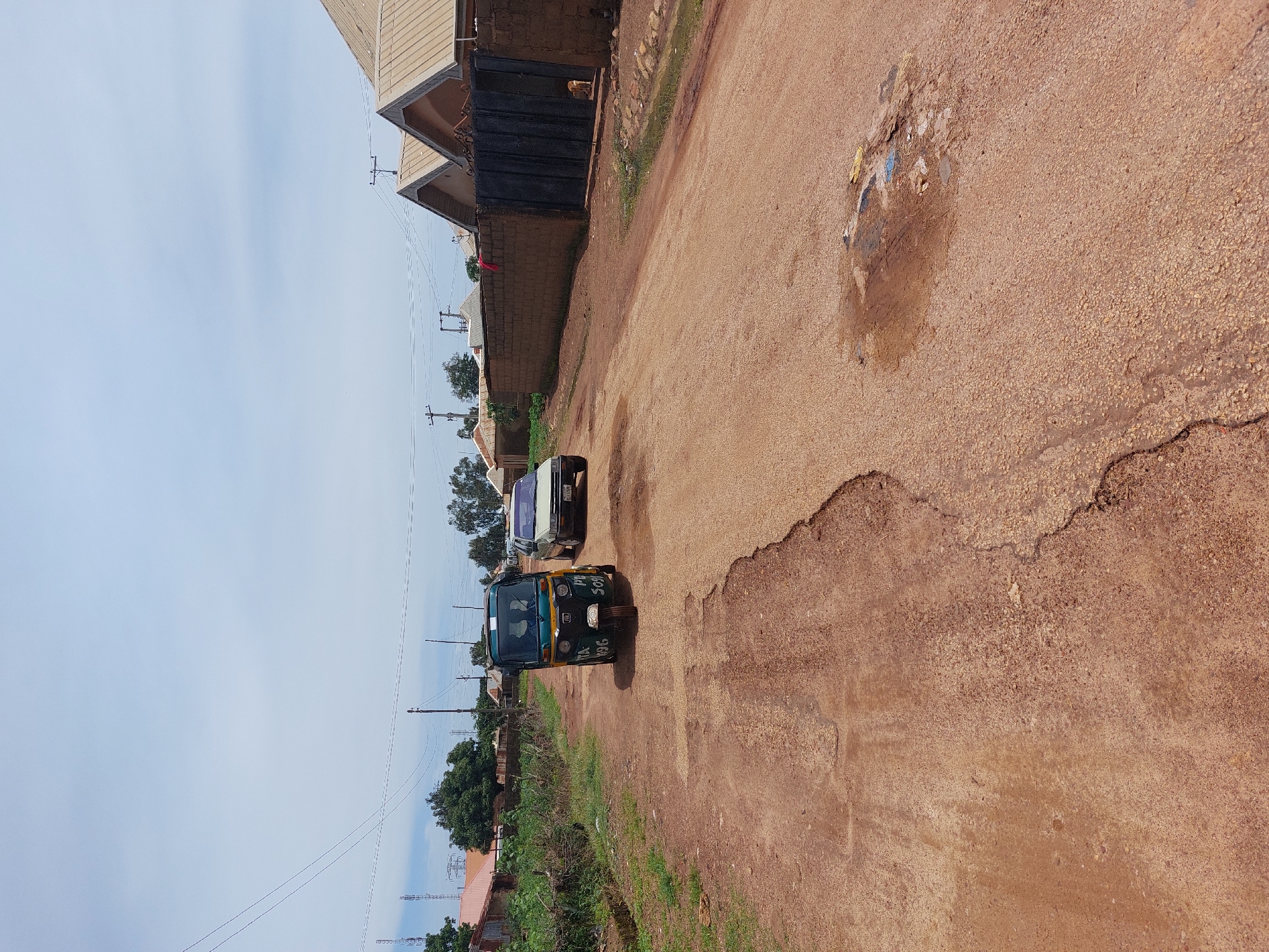 The image depicts a street scene in Barakin, Nigeria, on a clear day. The perspective is from a slightly elevated position, looking down a reddish-brown dirt road.
The primary subjects are three vehicles. In the foreground, a dark green and yellow auto-rickshaw is parked, with two individuals visible inside. Behind it, a white car is parked, followed by a light-colored sedan or SUV. The road itself is uneven and shows signs of wear, with a large pothole or eroded section in the right foreground. Debris, including colorful pieces of plastic and some wetness, is visible in a patch on the road.
To the left of the road, there are buildings with corrugated metal roofs and brick walls. A tall black metal gate is prominent, with a brick wall extending from it. Vegetation, including grass and trees, lines the side of the road, with some taller trees further in the background. Power lines and utility poles are visible against the pale blue sky, suggesting an inhabited area.
The overall impression is of a typical rural or semi-urban street in Nigeria. The time of day appears to be daytime, likely late morning or early afternoon, given the brightness of the sky and the shadows cast. The weather seems to be fair, with a clear sky. There is no obvious emotional tone conveyed by the scene; it is a straightforward depiction of everyday life.
Visible text includes markings on the auto-rickshaw. On the front, the number "986" is visible, along with other text that is difficult to decipher clearly but appears to include abbreviations like "MAX" and potentially names or route information.