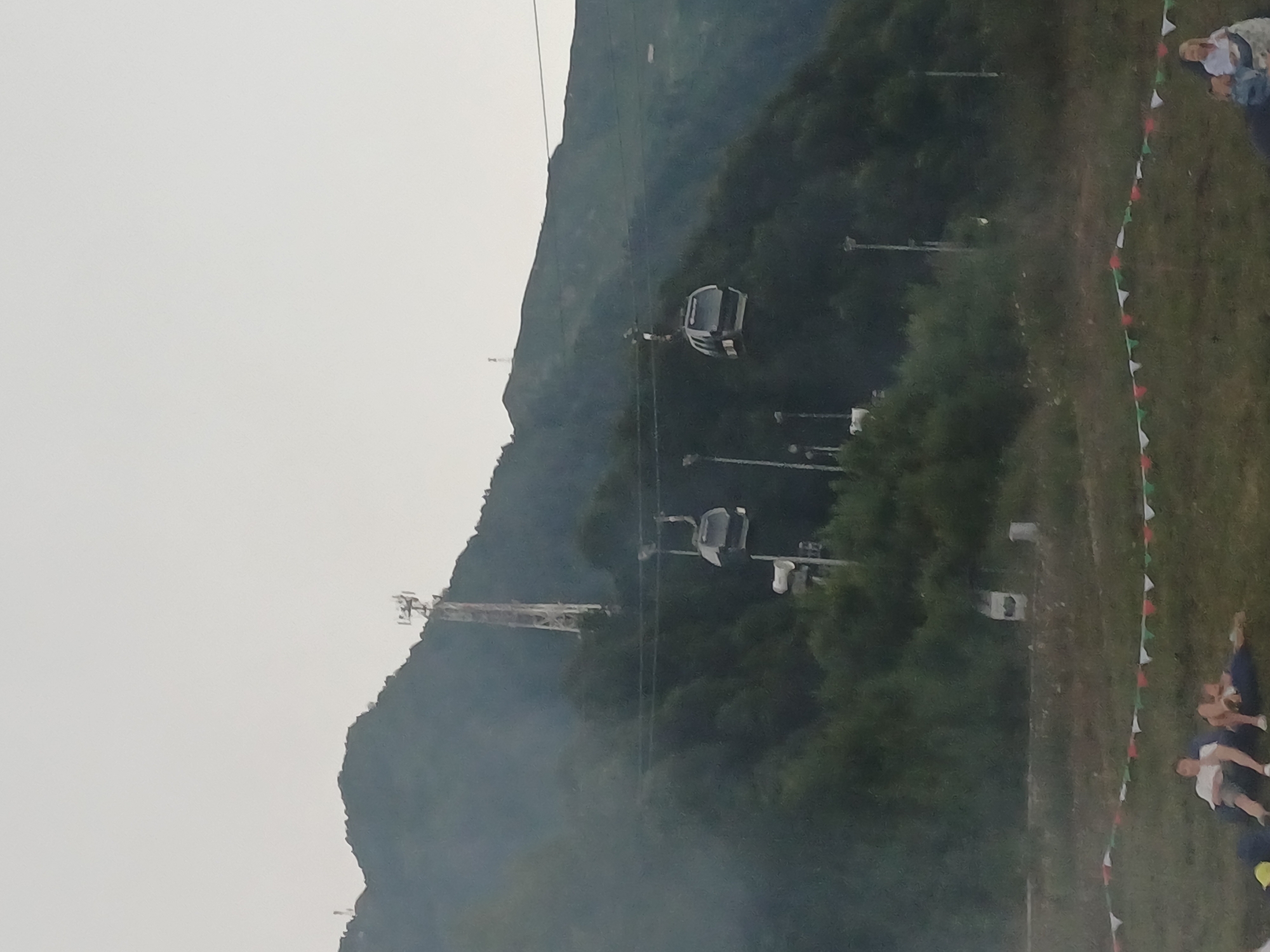 This image depicts a mountainous landscape, likely near the city of Estosadok, Russia. In the background, a tall, lattice-like tower, possibly a telecommunications mast, stands on a treed hillside. Two cable cars are visible suspended on wires, traversing the mountain slopes. The foreground features a grassy area with a string of decorative pennant flags in red, white, and green. Several people are seated or lying on beanbag chairs in the grass, appearing to relax and observe the scenery. The overall atmosphere suggests a recreational or tourist area, possibly a ski resort in the off-season or a mountain park. The lighting indicates it is either early morning or late afternoon, with overcast skies.