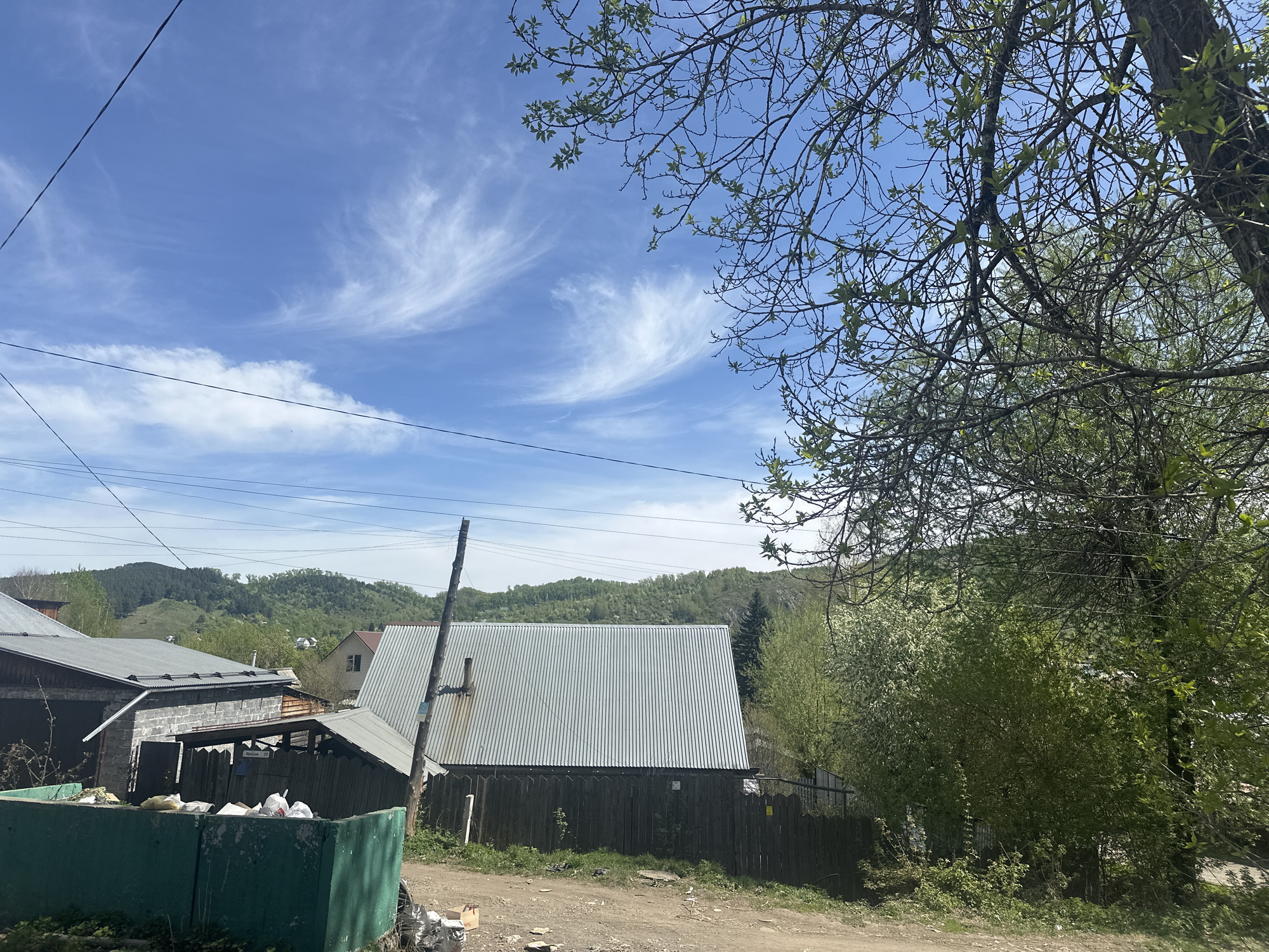 The image shows a street scene in Gorno-Altaysk, Russia. A dirt road runs along the bottom of the frame, with various trash items scattered on the ground. On the left is a green dumpster overflowing with white bags, likely containing refuse. A wooden fence and metal roof are visible in the midground. In the background, rolling hills covered with green vegetation are visible under a partly cloudy blue sky. A telephone pole and wires stretch across the top portion of the frame, adding to the cluttered aesthetic. A tree with new leaves is in the right corner of the frame.