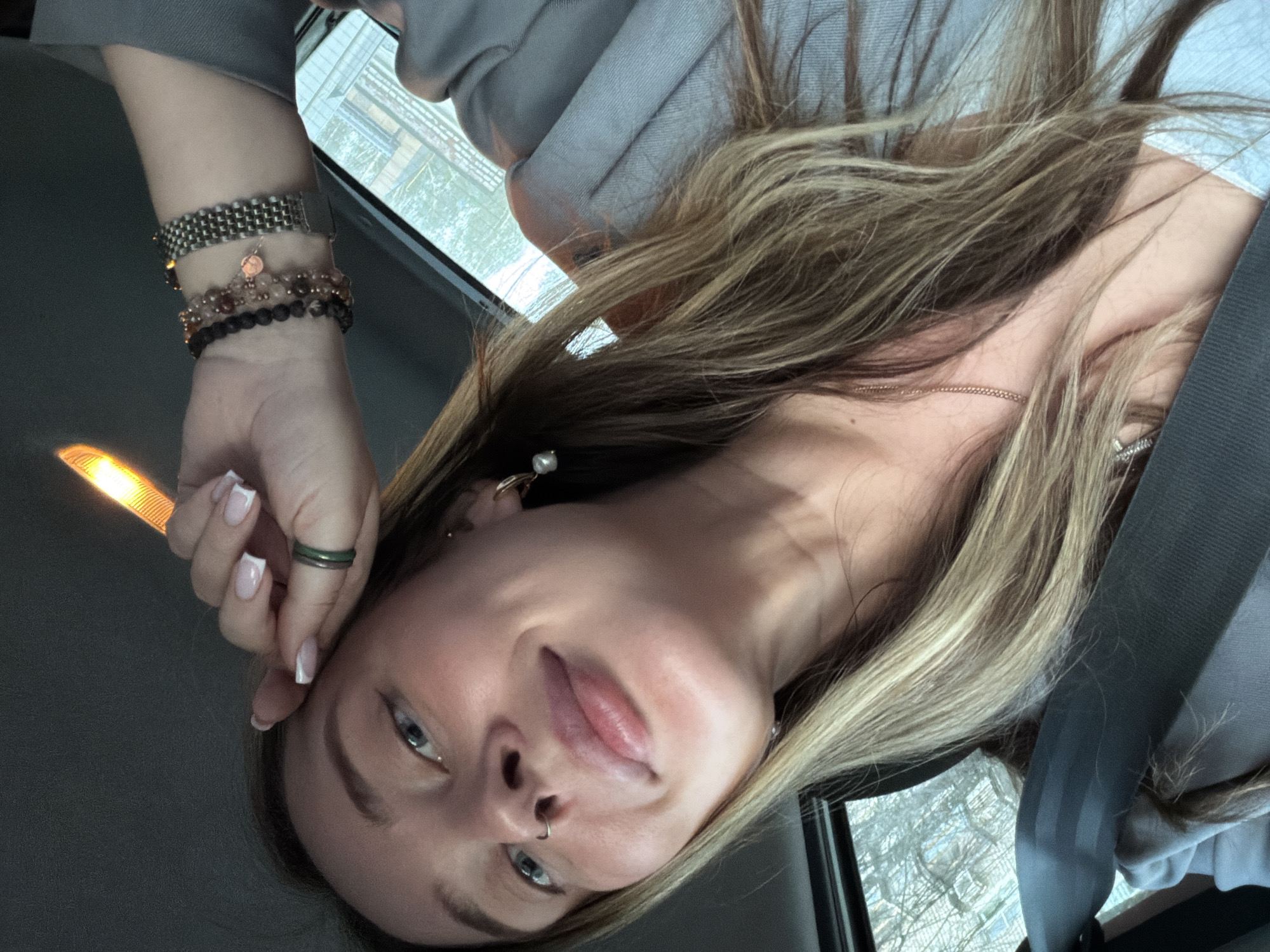 A medium close-up, eye-level shot of a young woman with blonde hair in a car. She's wearing a seatbelt. Her manicured hand is up near her face, adorned with a green ring, bracelets, and a silver watch. She appears to have a nose piercing. The location cannot be determined as Chaikovsky, Russia.