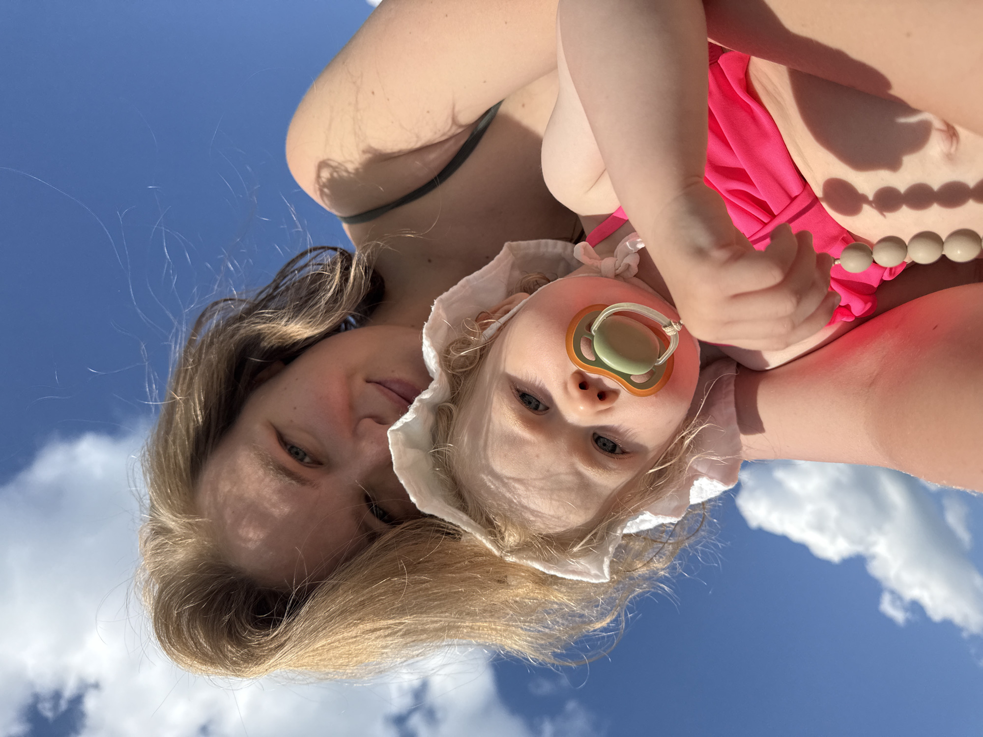 In this close-up, low-angle shot, a woman and a baby are positioned against a backdrop of a bright blue sky with scattered white clouds. The woman, with blonde hair, is looking down at the baby. The baby, wearing a white bonnet and a pink swimsuit, has a pacifier in its mouth and a string of wooden beads draped over its hand. The woman is wearing a dark green swimsuit top. The perspective suggests they are outdoors, possibly at a beach or park, given the bright sunlight and open sky. The location is not identifiable within the city of Колокша, Russia from the visual information provided.