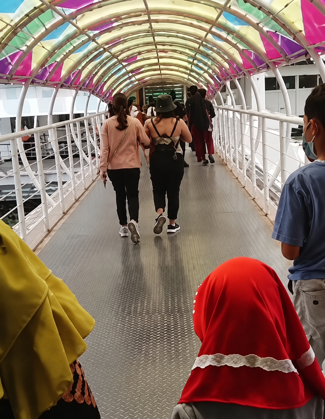 In a pedestrian walkway under a colorful, arched canopy, a group of individuals walk away from the camera. Two women in the foreground are walking side-by-side. The woman on the left wears a light pink long-sleeved shirt and black pants. The woman on the right wears a dark top with a pattern across her back, dark pants, and a dark green bucket hat. Behind them, several other people are visible walking in the same direction. On the right side of the walkway, a person wearing a blue t-shirt and a face mask is partially visible. In the immediate foreground, two individuals, one wearing a bright yellow garment and the other a red hijab with white trim, are also present. The walkway has a metal floor with a diamond plate pattern. The scene appears to be in a public area, possibly a terminal or a covered market. The location is identified as Berastagi, Indonesia.