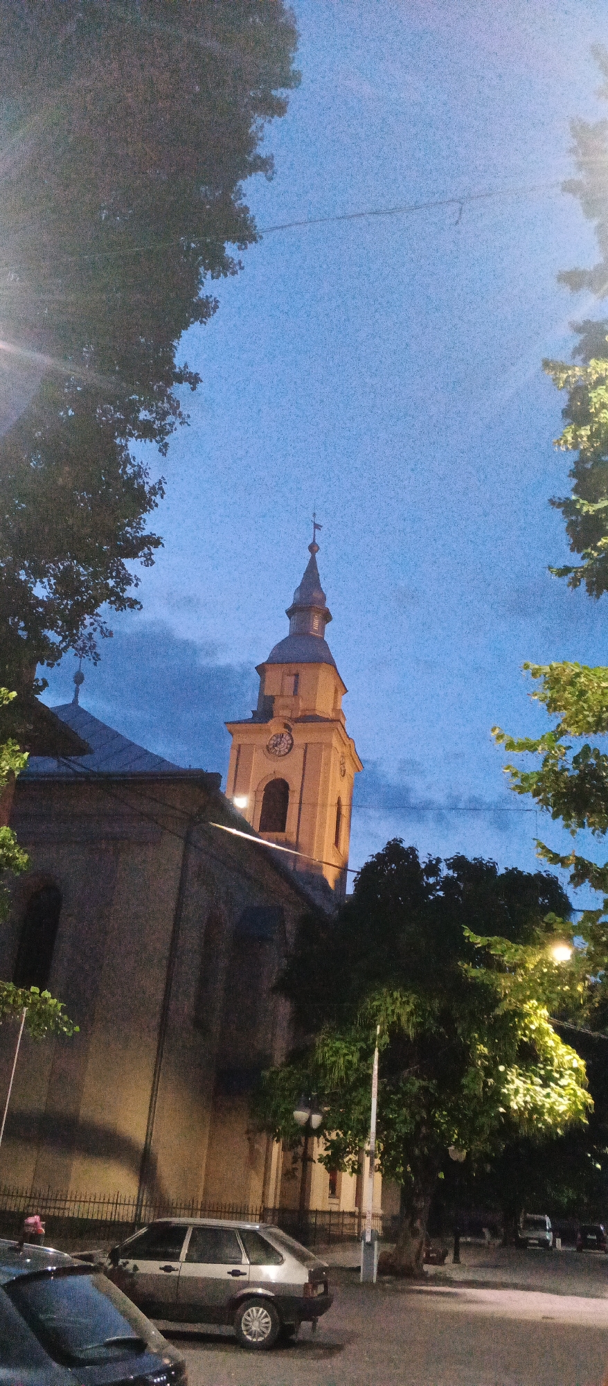 This vertical, low-angle photograph captures the bell tower and part of a church building in the city of Berehove, Ukraine, during twilight or early evening. The sky is a deep blue, transitioning to darker hues near the horizon, with some scattered clouds. A prominent yellow bell tower with a dark spire topped with a cross stands to the right of the frame, featuring a clock face with Roman numerals and visible clock hands. Adjacent to the tower is the side of the church, constructed with grey stone and illuminated by artificial light, casting shadows. Trees with lush green foliage flank the scene on both sides, with some branches extending into the foreground. The lower portion of the image reveals a street scene with several vehicles parked. In the foreground, a silver hatchback car is parked, and the rear of a dark-colored vehicle is visible to the left. Further back, other cars and streetlights are discernible. The overall atmosphere is tranquil, with the artificial lighting creating a warm contrast against the twilight sky.