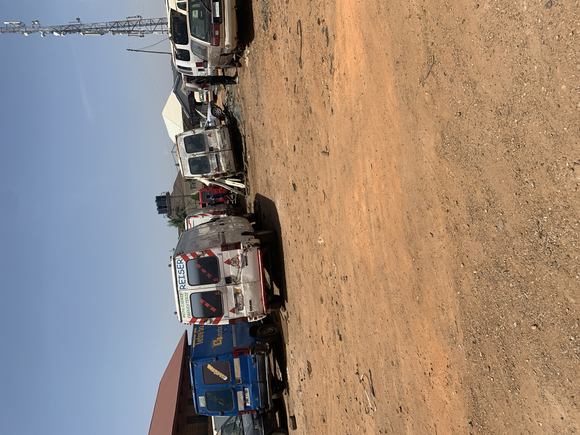 The image depicts an outdoor scene, likely a salvage yard in Rafin Par, Nigeria, showing a row of derelict vehicles. The foreground is dominated by a dirt ground. The vehicles appear to be predominantly vans, possibly service or delivery vehicles. Prominent markings include "REISER" and "letrebo" on some vans, suggesting European origins. A tall metal structure resembling a communication tower is visible in the background. The overall scene indicates an area where old vehicles are stored or scrapped. The sky is clear and blue.
