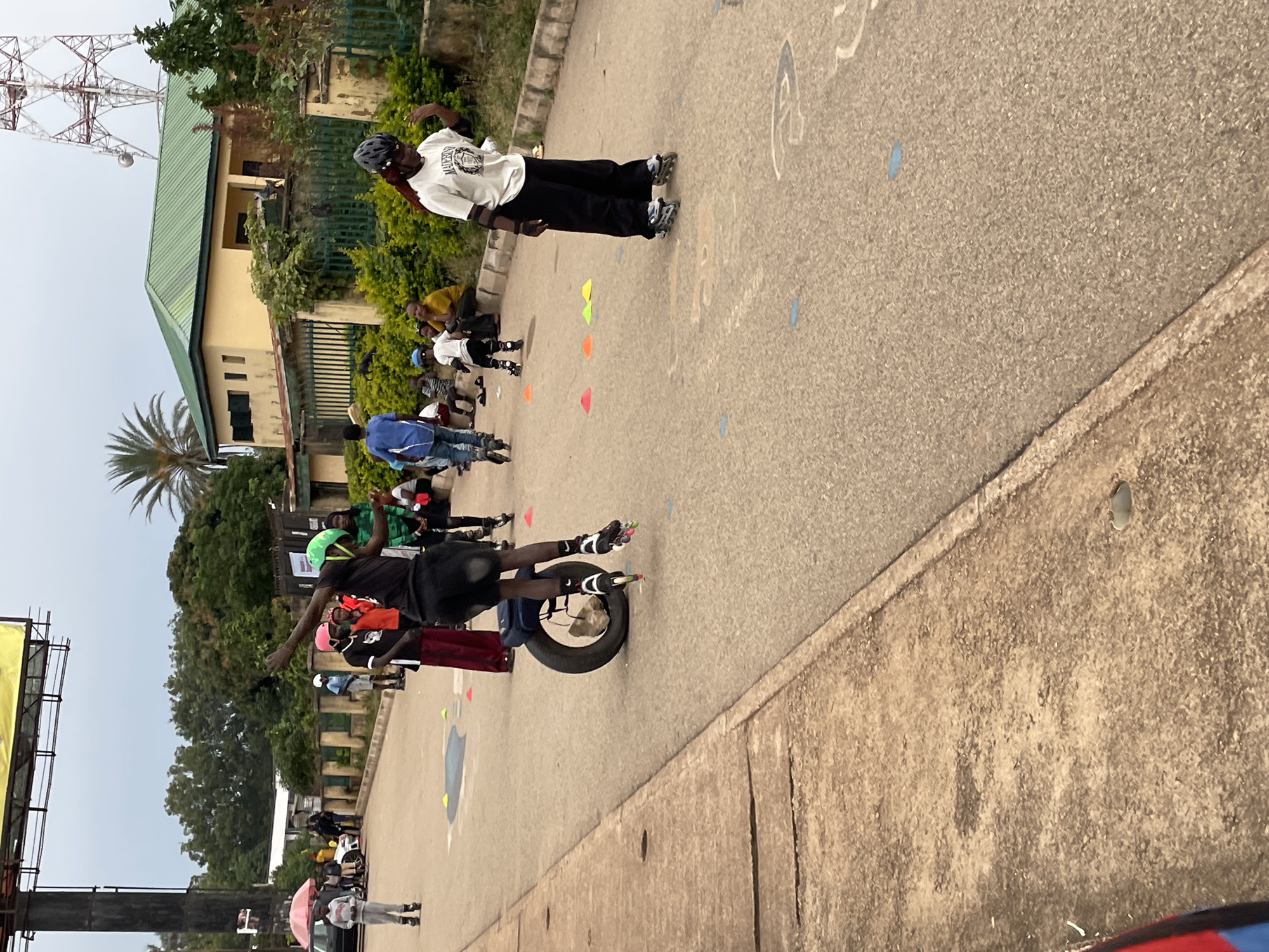 The image depicts an outdoor scene, likely a street or open paved area, in Jos, Nigeria. A group of people are engaged in inline skating. In the foreground, a person wearing a green helmet and dark clothing is captured mid-action on rollerblades, with one leg extended. To the right, another individual in a white t-shirt and black pants, also on rollerblades, is visible from the waist down, with their right hand raised.
In the middle ground, several other individuals are present. Some are seated on a curb, while others stand and observe. A few children are also seen in rollerblades, navigating the paved area. Cones, in various colors such as orange, yellow, and blue, are scattered on the ground, suggesting a designated area for skating or an obstacle course.
The background reveals a building with a green roof, characteristic of architecture in the region. A prominent telecommunications tower stands tall in the distance, a common landmark in urban Nigerian landscapes. The overall environment appears to be a community gathering or an organized skating event taking place in a somewhat urbanized yet open setting within Jos. The weather appears to be overcast.