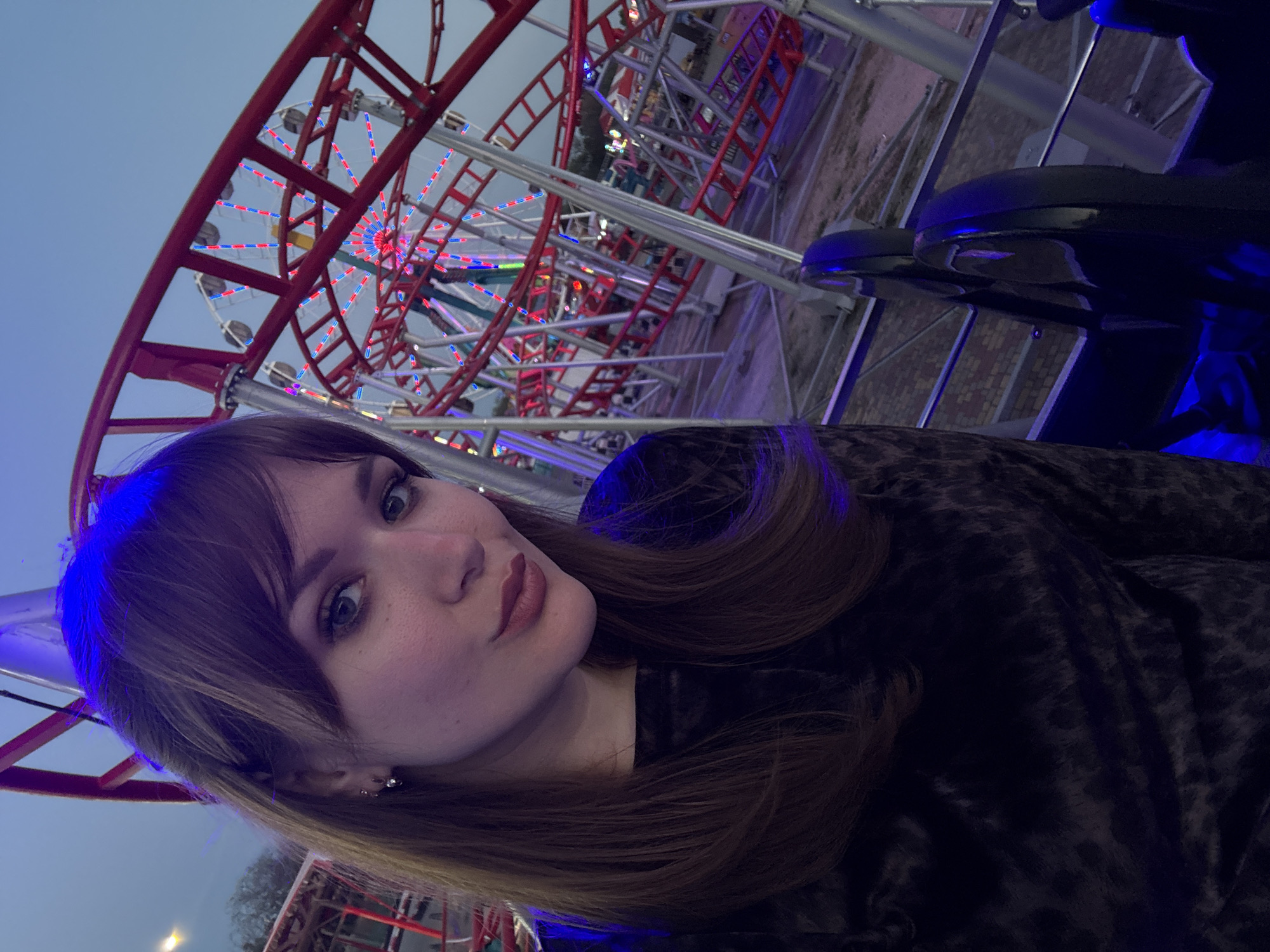 The image is a selfie of a fair-skinned woman with shoulder-length brown hair, wearing a black animal-print jacket. She has bangs and is looking directly at the camera with a slightly pursed expression.
The background shows an amusement park or fairground. The most prominent feature is a roller coaster with red and white tracks. There's also a Ferris wheel visible in the background, decorated with red, white, and blue lights. The roller coaster's metal framework and various structures are visible around the woman. The image has a blue tint. The location is likely a fairground or amusement park. Without any further information or clues, I can't confirm this image was taken in Yevpatoriya, Ukraine.