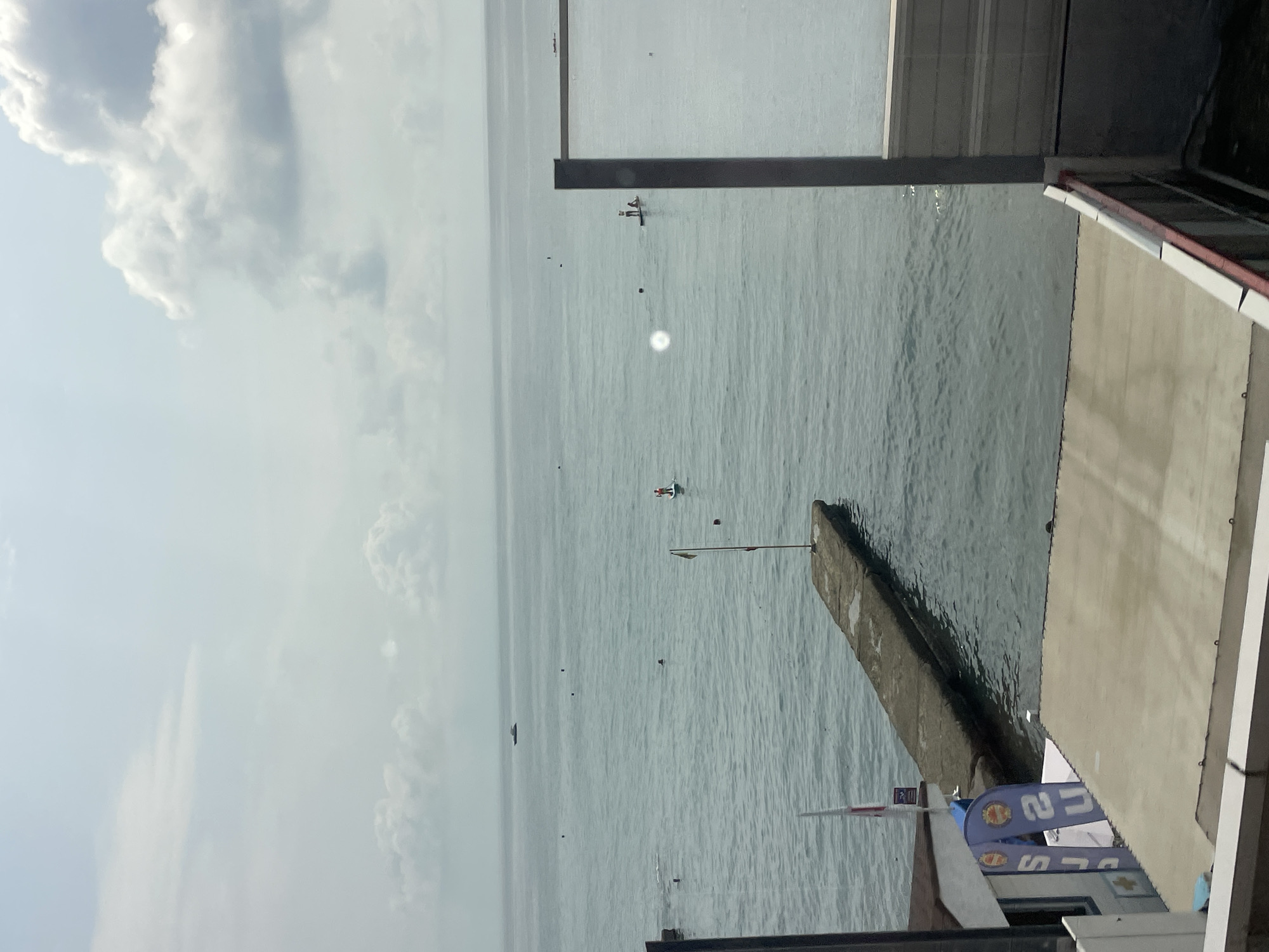 The image depicts a view of the Black Sea from a balcony or elevated vantage point in Sochi, Russia. In the foreground, there are parts of a building and a deck with a railing. The middle ground shows a concrete pier extending into the water, with a flag on a pole. The sea itself is calm with gentle ripples. Several people are visible in the water: some are swimming, and others are paddleboarding. In the distance, a small boat can be seen on the horizon. The sky is partly cloudy. The overall impression is of a sunny day at a seaside resort.