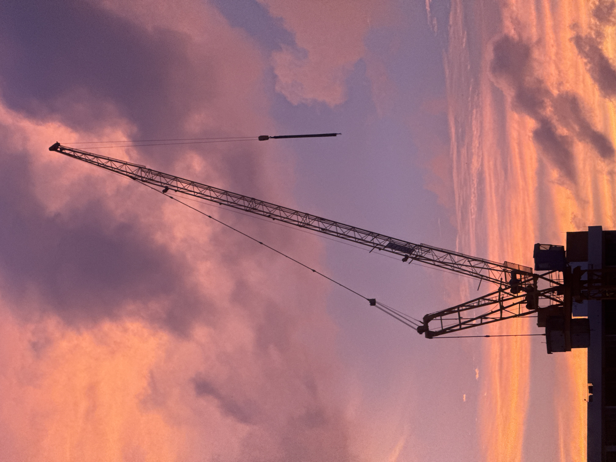 A silhouette of a construction crane stands against a vibrant sunset sky in Karon, Thailand. The sky is filled with dramatic clouds painted in hues of orange, pink, and purple. The crane, a tall, skeletal structure, dominates the frame, its long boom extending diagonally upwards. Part of a building is visible at the bottom of the image, providing a base for the crane. The scene is tranquil, with no people or animals present, suggesting a pause in construction activities. The overall impression is one of industrial might juxtaposed with the ephemeral beauty of a tropical sunset.
