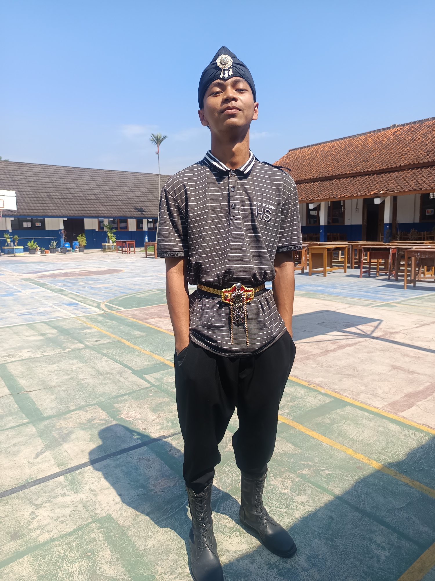 A young male stands outdoors on a paved surface, possibly a schoolyard, on a bright, sunny day. He is looking directly at the camera and appears to be in his late teens or early twenties. He is wearing a traditional black headpiece adorned with a silver emblem, a striped polo shirt with "HS" embroidered on the left chest, black pants, and black lace-up boots. A decorative gold and red belt with a large, ornate buckle is worn around his waist. The background features buildings with tiled roofs and what appears to be a basketball court with painted lines. The location is identified as Tanjungsari, Indonesia. The scene suggests a formal or ceremonial occasion, possibly related to a school event or cultural gathering.