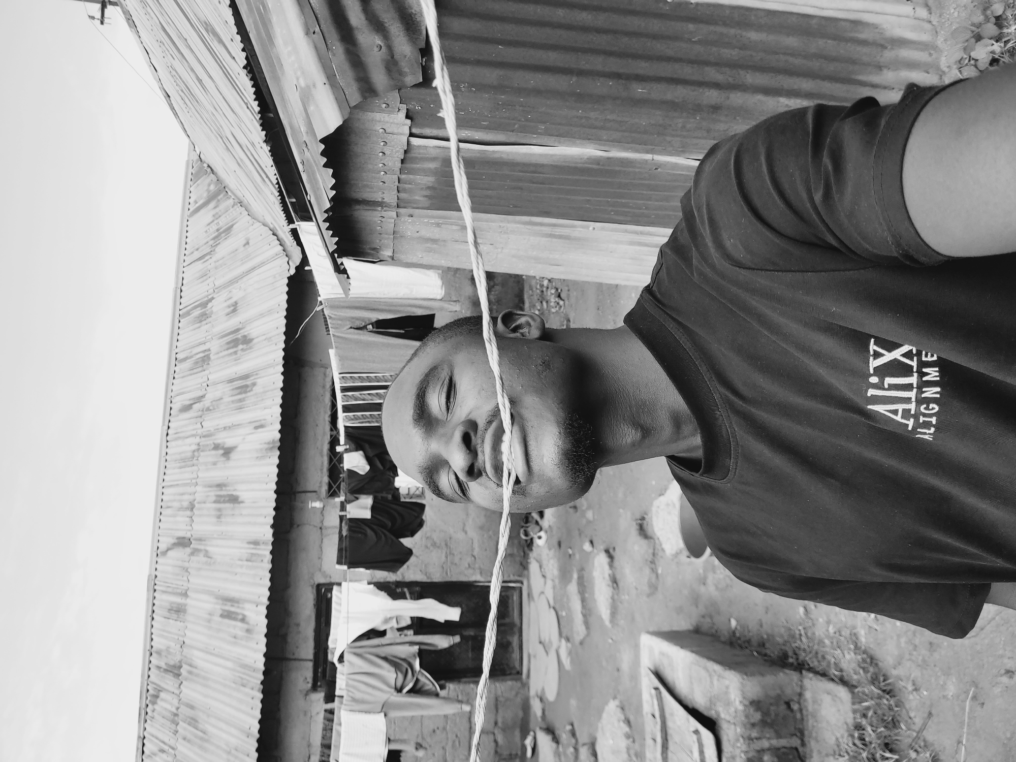 The black and white image depicts a dark-skinned man standing outside in what appears to be a residential area in Bukuru, Nigeria. He is facing the camera, with a piece of rope positioned between his lips and nose. The man is wearing a dark t-shirt with white lettering on the chest, part of which reads "ALIGNME". Behind him, there is a corrugated metal structure that could be a house or shed, with clothes hanging on a line strung in front of it. The ground is uneven and partially covered in vegetation. A concrete structure is visible in the lower right corner.