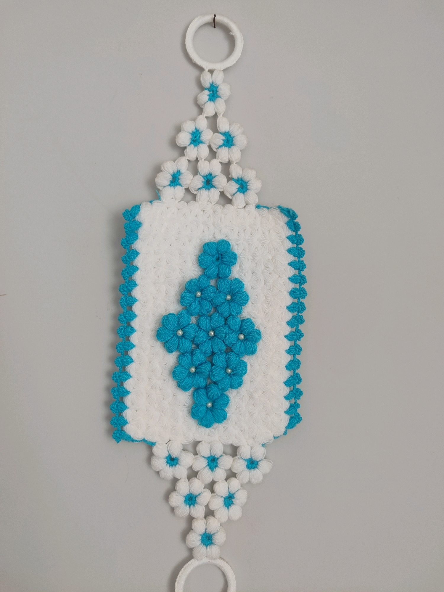 A white knitted hanging decoration with blue floral accents is displayed against a plain grey wall. The decoration features a rectangular central panel adorned with a diamond pattern of blue crocheted flowers, each with a small pearl in the center. The edges of the central panel are trimmed with a blue crocheted border. Extending from the top and bottom of the central panel are chains of smaller white and blue flowers, culminating in white rings at the top and bottom for hanging. The lighting is even, and the image is taken from a straight-on perspective, focusing on the decorative item. There are no people or indications of location within a city.