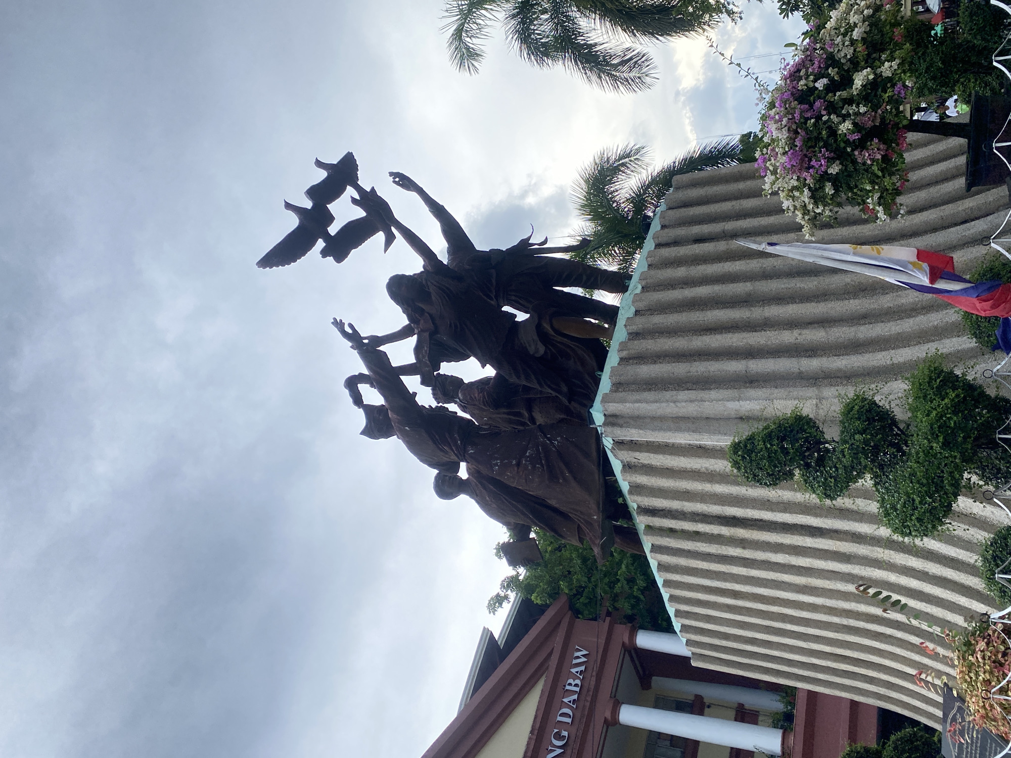The image shows the Rizal monument in Rizal Park, Davao City, Philippines. The monument is a bronze sculpture depicting Jose Rizal and other figures, with doves flying above. The monument is situated on a concrete base with vertical ridges. A building with the text "DABAW" is visible in the background. Palm trees and flowering bushes surround the monument. A Philippine flag is also present. The sky is overcast.
