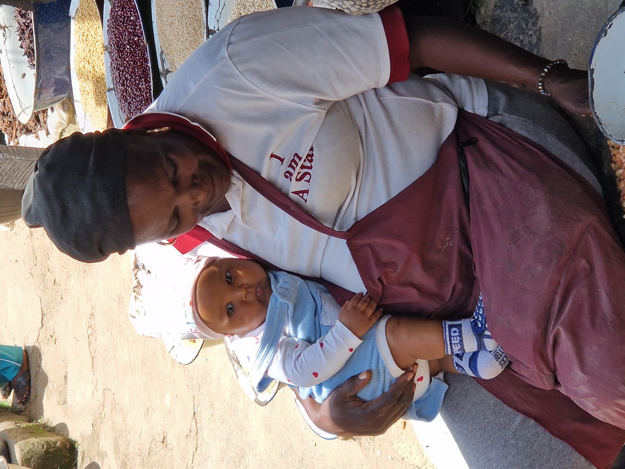 The image depicts a woman holding a baby in what appears to be an outdoor market setting, possibly in Kabong, Nigeria. The woman, wearing a white polo shirt with red trim and a dark head covering, is looking down, while the baby, dressed in a blue romper and a pink cap, looks directly at the camera. The baby is wearing white socks with blue stripes and the word "SPEED" printed on them. The background shows large metal bowls filled with various grains or seeds, indicative of a market stall. The ground is made of packed earth, and a partial view of a person's feet wearing brown sandals and blue trousers is visible in the upper left corner, suggesting a bustling environment. The lighting suggests it is daytime.