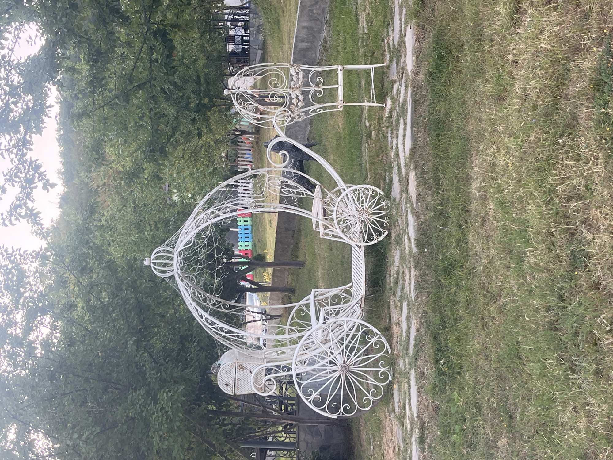 In Ismailli District, Azerbaijan, a white wrought iron carriage-shaped bench sits on a paved area surrounded by grass. The carriage features intricate scrollwork and a decorative canopy. Behind the carriage, a dark statue resembling a dog or wolf is visible. In the background, trees line the property, and a colorful wooden fence encloses a playground or seating area. A concrete barrier separates the grass from another area with tables and chairs, suggesting a restaurant or outdoor venue. The lighting indicates it is daytime, likely late afternoon.