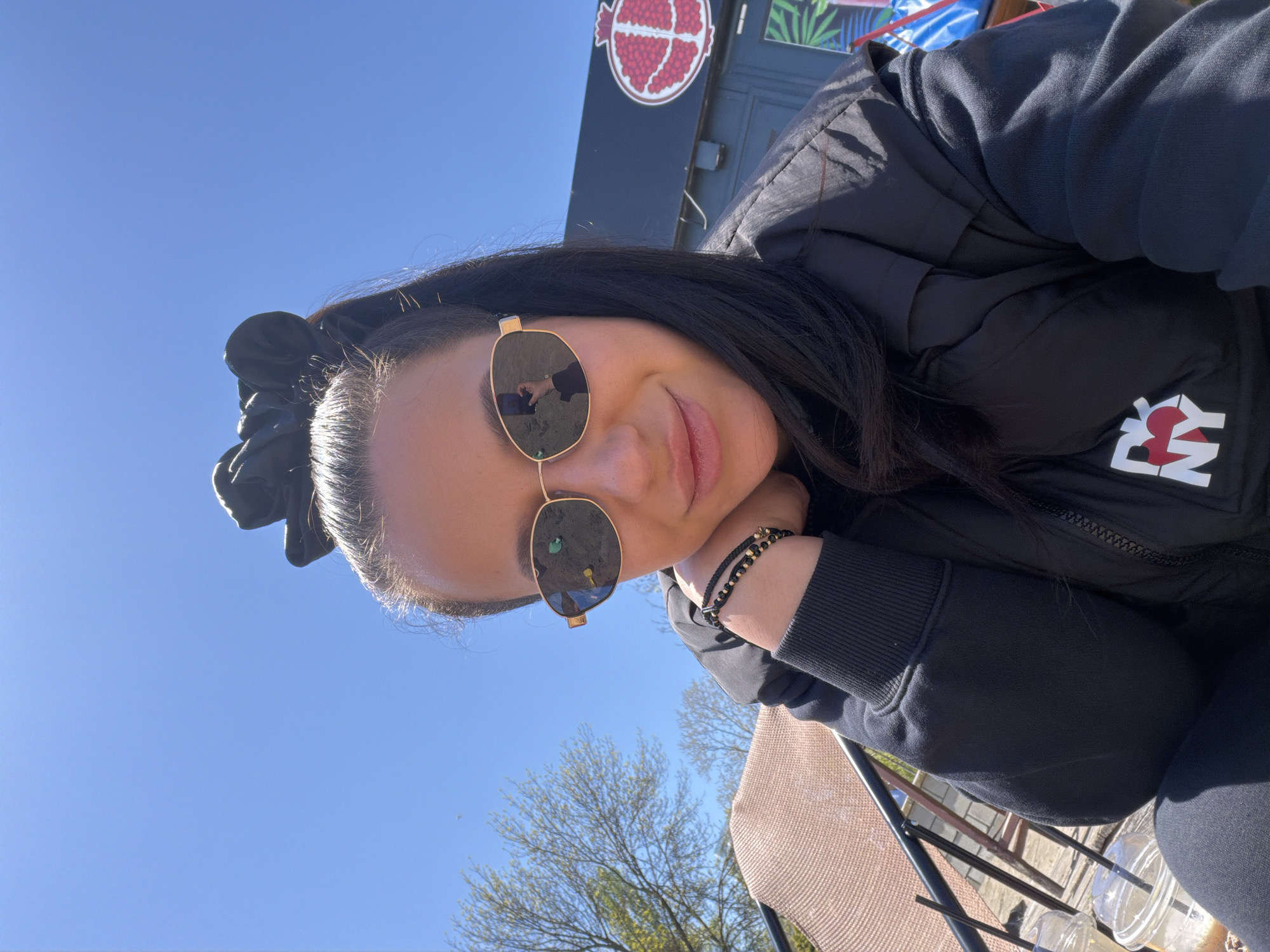 A woman is outdoors in an unknown location in the city of Unknown. She is wearing sunglasses, a black jacket with a "NY" logo on the front, and a black scrunchie in her hair. She is resting her head on her hand, which has a black beaded bracelet on the wrist. A chair and a plastic cup with straws are visible in the foreground. A building with a red circular logo and a blue sky with trees are in the background.