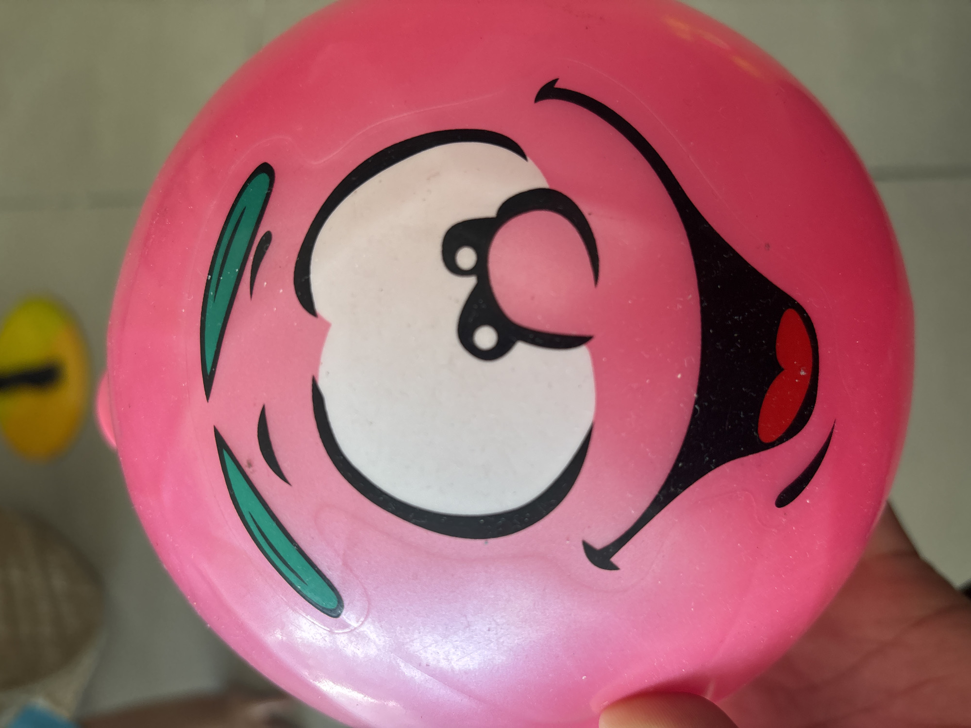 A close-up, eye-level shot captures a pink, inflatable ball with a cartoonish face. The face features large white eyes with black pupils, green arched eyebrows, and a wide, red-lipped smile. A black nose with two small circles completes the playful expression. The ball is being held by a hand visible in the lower left corner of the frame. The background is blurred, showing hints of other objects, one of which appears to be a yellow circular item. The lighting suggests an indoor or shaded outdoor setting. There is no visible indication of the specific location within Dubai, United Arab Emirates.