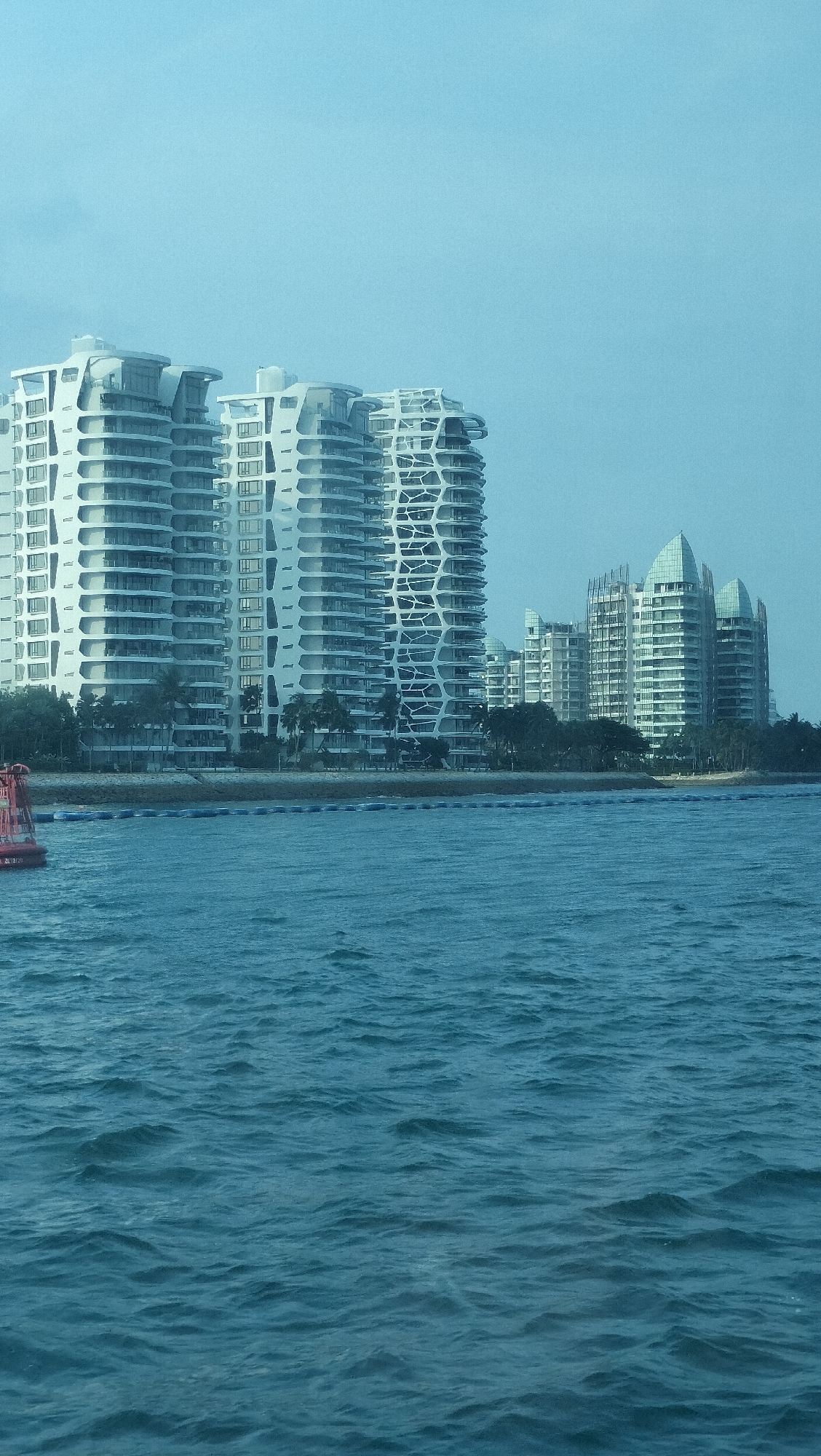 The image shows a waterfront view of modern residential buildings in Singapore. In the foreground, the turquoise water of the sea is visible with gentle waves. A red and white buoy is partially visible on the left side of the frame. In the background, a series of tall, white, curvilinear apartment buildings with many balconies and windows are lined up along the coast. Behind these buildings, other apartment complexes with a more conventional, geometric design and sloped roofs are visible. Lush green trees, likely palm trees, are interspersed between the buildings and along the coastline. The sky is a pale, hazy blue. This scene depicts a residential area along the coast of Singapore, likely a developed urban waterfront.