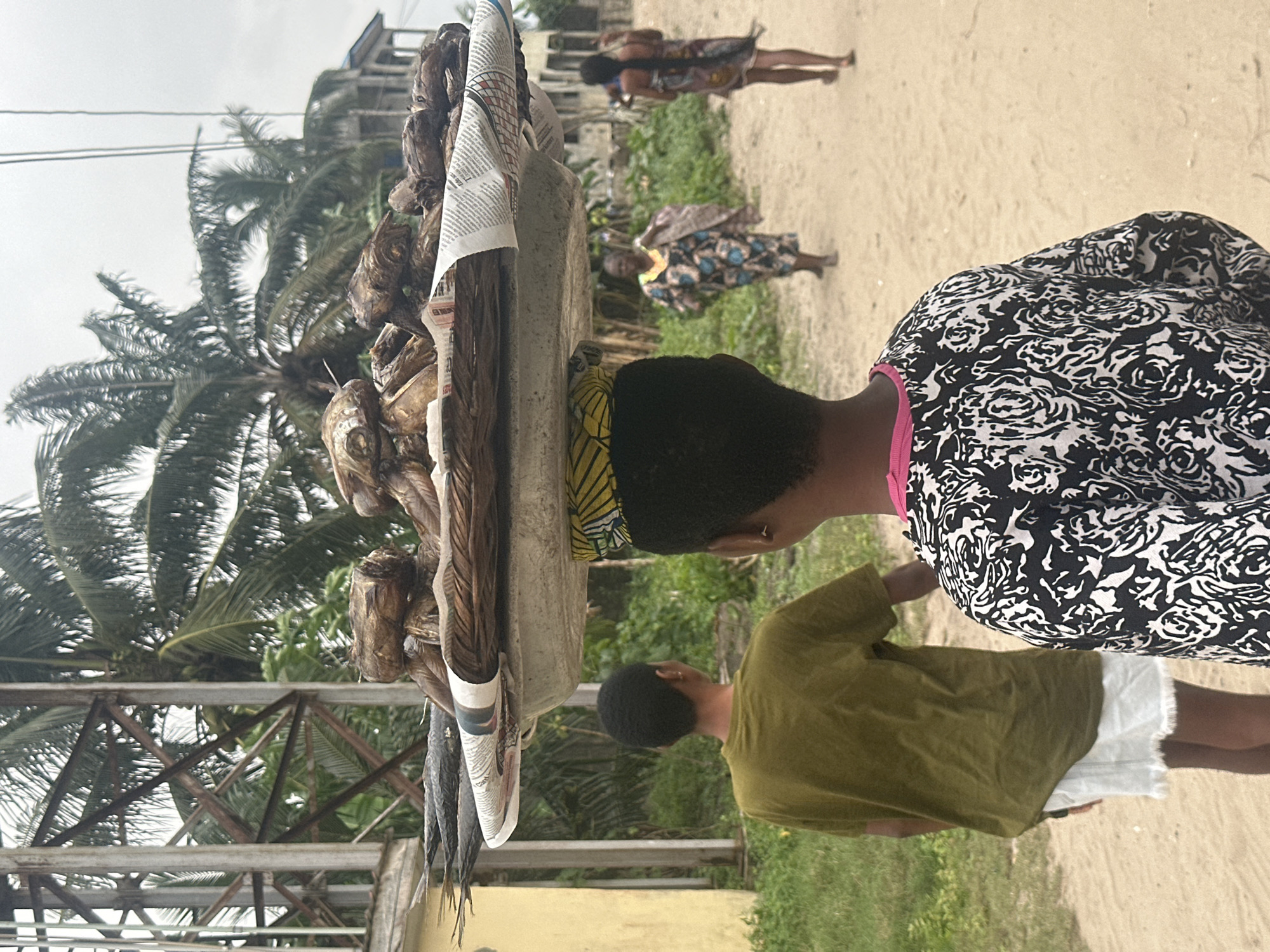 An outdoor, medium shot shows a young woman carrying a large tray of dried fish on her head, walking on a dirt road. She is wearing a black and white patterned shirt and a colorful headwrap. In front of her, a person wearing a green t-shirt and white shorts walks in the same direction. Further down the road, two women are visible, one in a patterned dress and the other in a patterned wrap. Palm trees and some buildings line the background, suggesting a tropical or coastal environment. The image appears to be taken in the city of Badagry, Nigeria.