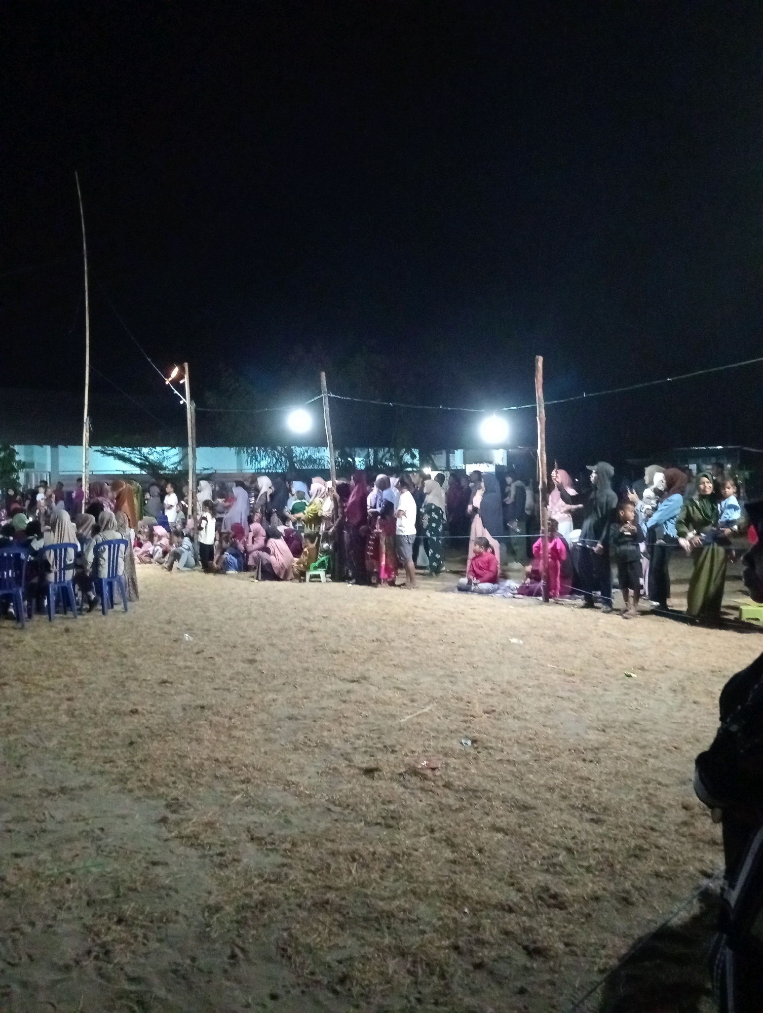 This image depicts an outdoor gathering at night in Pagerungan Kecil, Indonesia. A crowd of people, dressed in a variety of attire including headscarves and traditional clothing, are congregated on a sandy, grassy area. Several wooden poles are visible, some supporting lighting fixtures that illuminate the scene. In the foreground, blue plastic chairs are arranged, suggesting a seating area. The background shows a building with windows and trees. The overall atmosphere appears to be festive or social, with individuals standing, sitting, and interacting in groups.