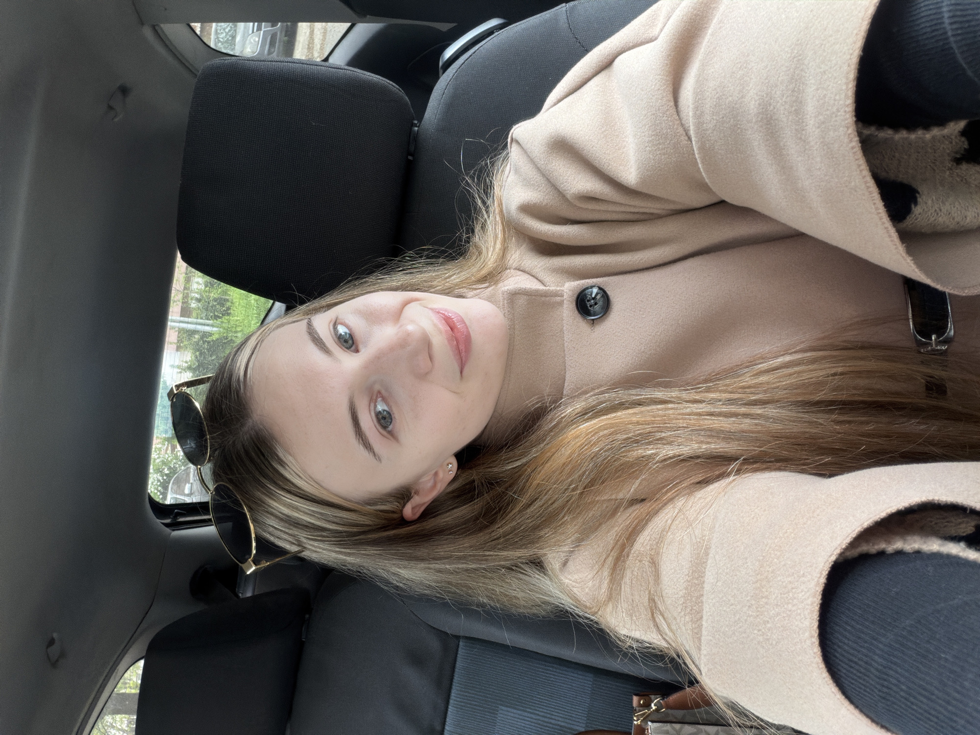 A blonde woman is photographed in the interior of a car. She is wearing a tan coat with one visible button. Sunglasses are tucked into her hair. The interior of the car is black. It is impossible to determine the location of the image in the city of Plovdiv, Bulgaria.