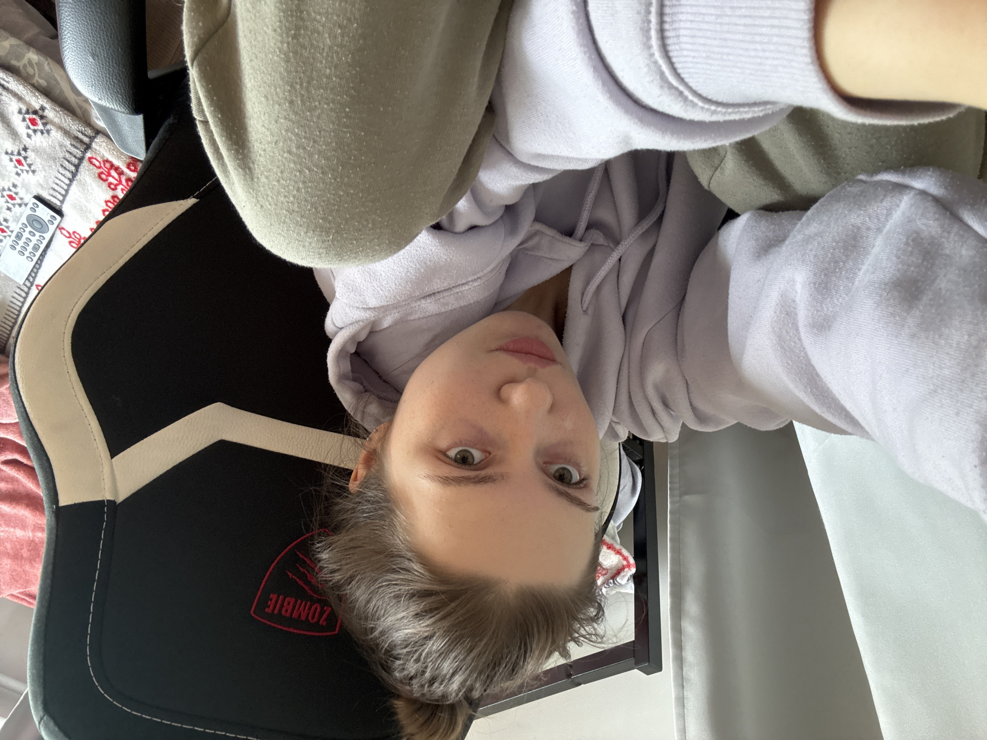 The image is a selfie of a person with fair skin, light brown hair pulled back in a low bun, and light-colored eyes. They are wearing a lavender hoodie and a green sweatshirt. The background includes a black and white gaming chair with a red "ZOMBIE" logo, a patterned blanket, and a white curtain. No information is available from the image to confirm the location in Saint Petersburg, Russia.