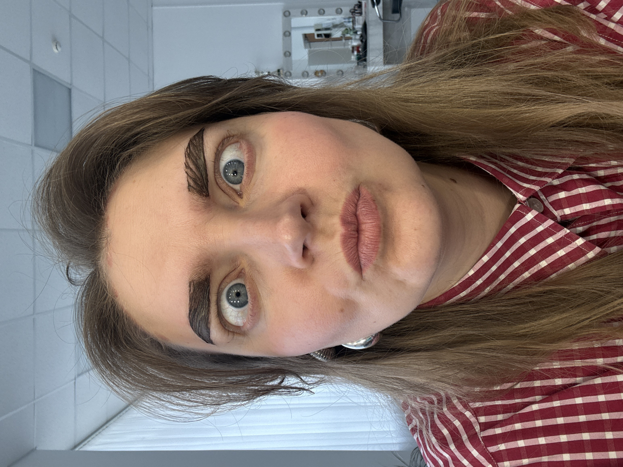 A close-up portrait captures a person, likely female, with long, light brown hair, gazing directly at the viewer with wide, light blue-grey eyes. The image is taken from a slightly low and rotated angle, presenting the subject's face prominently.
The most distinctive feature is the person's eyebrows, which are heavily and artfully made up. They are thick and dark, styled with visible, parallel strokes that create a feathered or ridged, almost segmented pattern. The wide-open eyes reflect circular lights, indicative of a vanity mirror. The person's lips are slightly parted and appear to be wearing a natural or nude-toned lipstick. Rosy blush is visible on the cheeks. A large, spherical, silver-toned earring can be seen on one earlobe.
The individual is wearing a red and white checkered button-up shirt. The background features light-colored tiled walls and a vanity mirror with lights, suggesting an indoor setting such as a salon, makeup studio, or dressing room. A horizontal blind or light-colored surface is also visible in the lower portion of the frame. The expression conveys a sense of directness, perhaps surprise or playful intensity. No visible text is present in the image. The location context provided is Izhevsk, Russia.