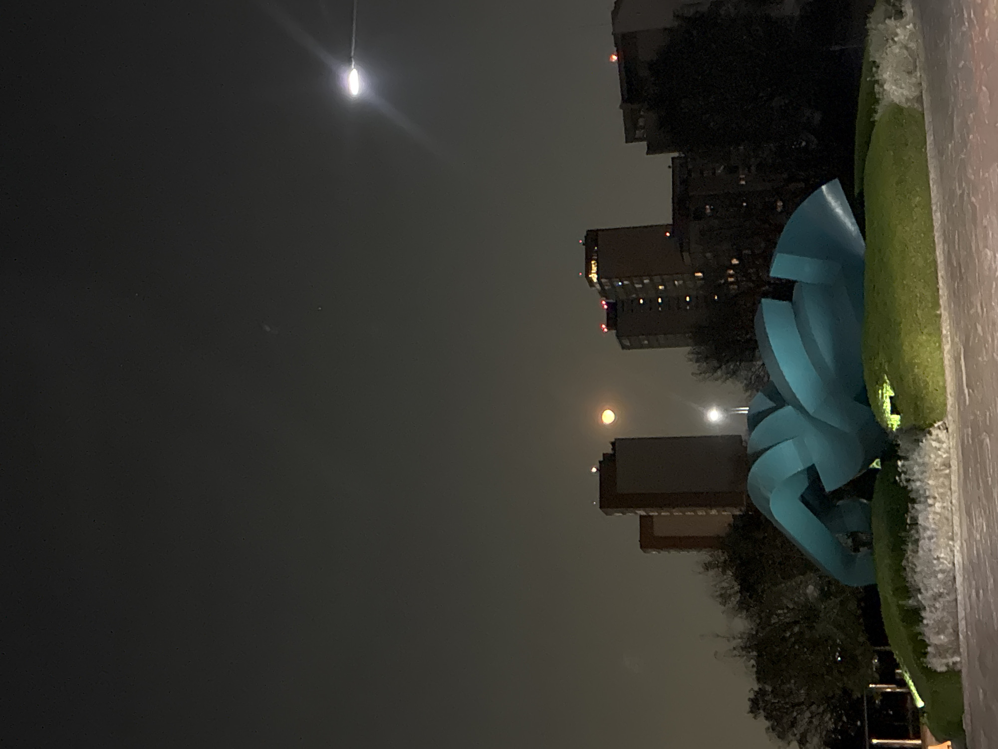 Here is a description of the image:
The image depicts an outdoor night scene in Huixquilucan de Degollado, Mexico. A blue, abstract sculpture sits in a green, landscaped area, possibly a garden or park. Buildings, including high-rises with illuminated windows, are visible in the background, indicating an urban setting. The sky is dark, with at least two bright lights—one at the top of the frame likely from a street lamp and another possibly from the moon lower in the image. The overall ambiance is dim, with artificial lights and the moon providing the primary illumination.