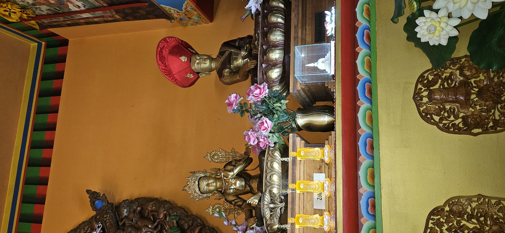 The image depicts the interior of a Buddhist temple in Zalaszántó, Hungary, specifically within the Béke Sztúpa (Stupa of Peace). The scene is dominated by a vibrant orange wall, adorned with a decorative border featuring alternating red, green, and blue stripes.

On a raised wooden shelf, two golden statues of Buddhist figures are prominently displayed. The statue on the left is seated, wearing an ornate crown and a golden robe, with its right hand raised in a gesture of blessing. The statue on the right is also seated and appears to be wearing a ceremonial red hat with golden embellishments. Both statues rest on elaborate lotus-shaped pedestals.

In front of the statues, a collection of objects is arranged. To the left of the statues, three small, translucent yellow figurines of standing Buddhist monks are positioned on individual lotus bases. A label reading "Zólu" is visible in front of the middle figurine. Between the statues, a vase filled with artificial pink roses and green leaves sits on the shelf. To the right of the vase, a small, white stupa is enclosed within a clear glass box. Beside the glass box, a white artificial lotus flower rests on the shelf, and to the far right, more artificial white lotus flowers and green leaves are visible.

Below the shelf, another decorative border is present, this one featuring a repeating pattern of colorful, stylized cloud-like shapes in red, blue, green, and yellow. The lower part of the image shows a yellow wall with intricately carved wooden panels featuring floral motifs and Buddhist symbols.