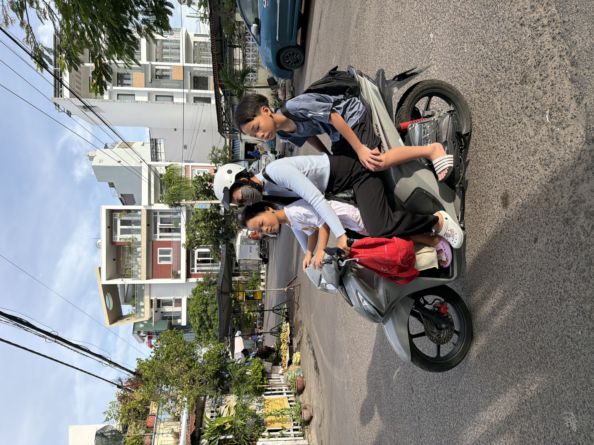 This is a media file of three people on a scooter in Nha Trang, Vietnam. The subjects are two children and one adult. The child in front is wearing a white shirt and black pants, and appears to be operating the scooter. The child in the middle is wearing a light blue shirt and black pants, and has a backpack on. The adult behind them is wearing a white helmet and a mask, and is holding onto the child in the middle. The scene is a street in a city, with buildings and trees visible in the background. The time of day appears to be daytime, and the weather is sunny. There is some text visible on the scooter, but it is too blurry to read. The overall mood of the image is neutral, with no strong emotions conveyed by the subjects.