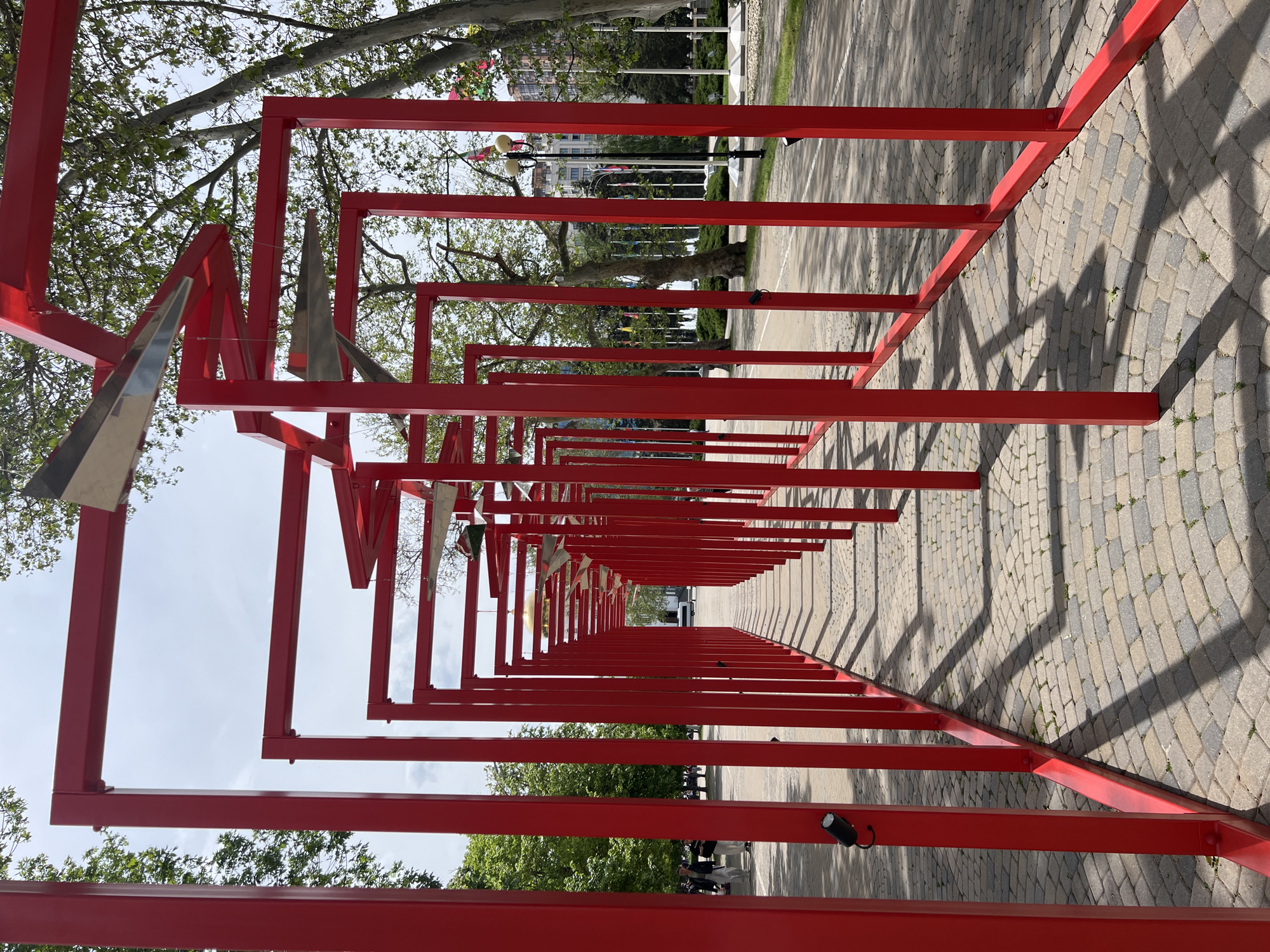 The image shows a red metal structure in a park, located in Krasnodar, Russia. The structure consists of a series of connected, diminishing red square frames that stretch out into the distance, creating a linear tunnel effect. Some of the frames are adorned with silver, triangular metallic shapes. The ground is paved with interlocking gray and brown stones. Trees line the sides, casting shadows on the pavement and the red metal. In the distance, beyond the structure, are trees, flag poles and a building. There are people visible in the distance to the right. The sky appears overcast.