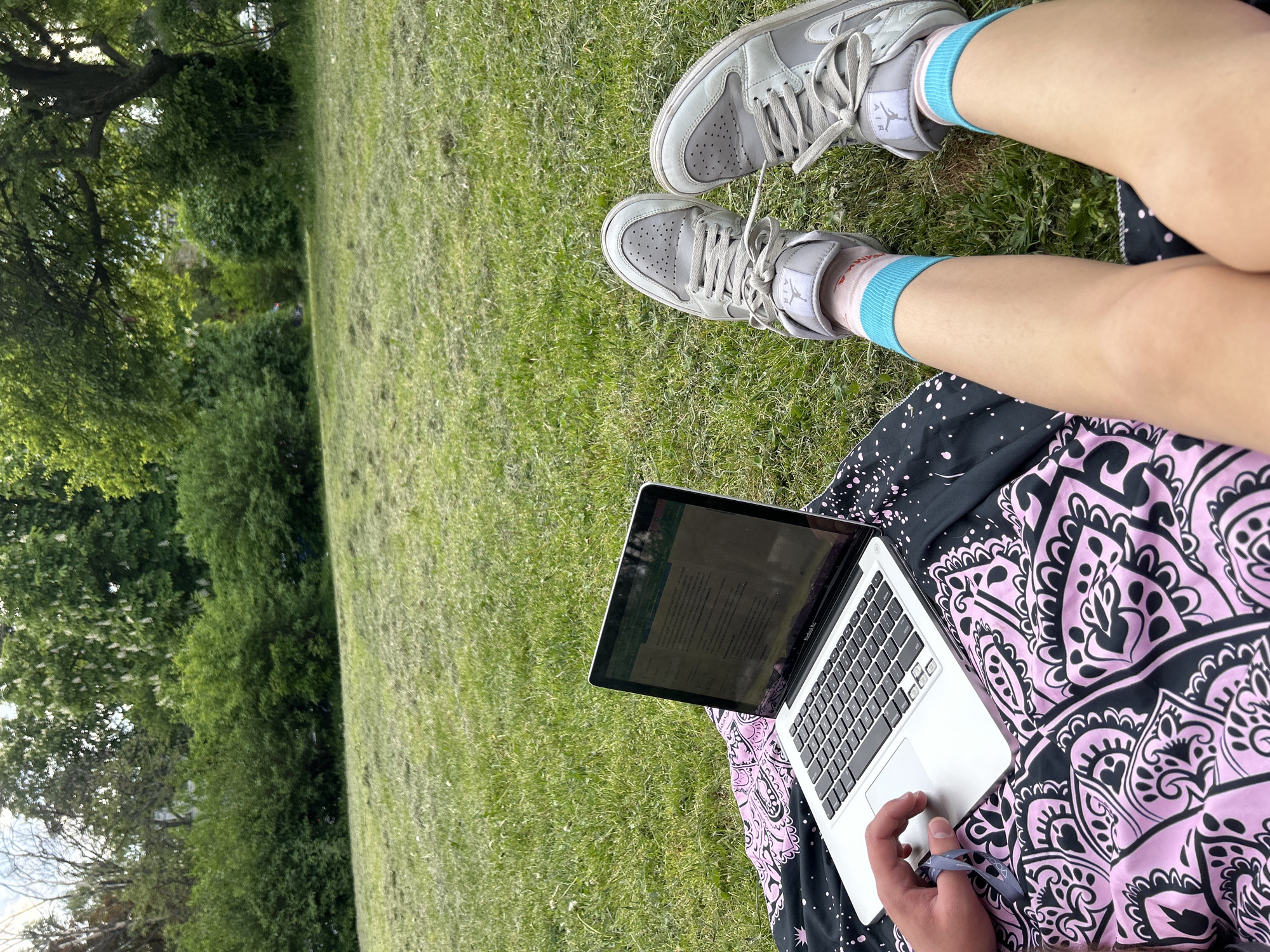 A person is sitting on a patterned blanket in a grassy area, using a MacBook laptop. The person is wearing gray sneakers with light blue socks. The grassy area is likely a park. There are trees in the background. The photo does not contain enough detail to identify the precise location in Sofia, Bulgaria.