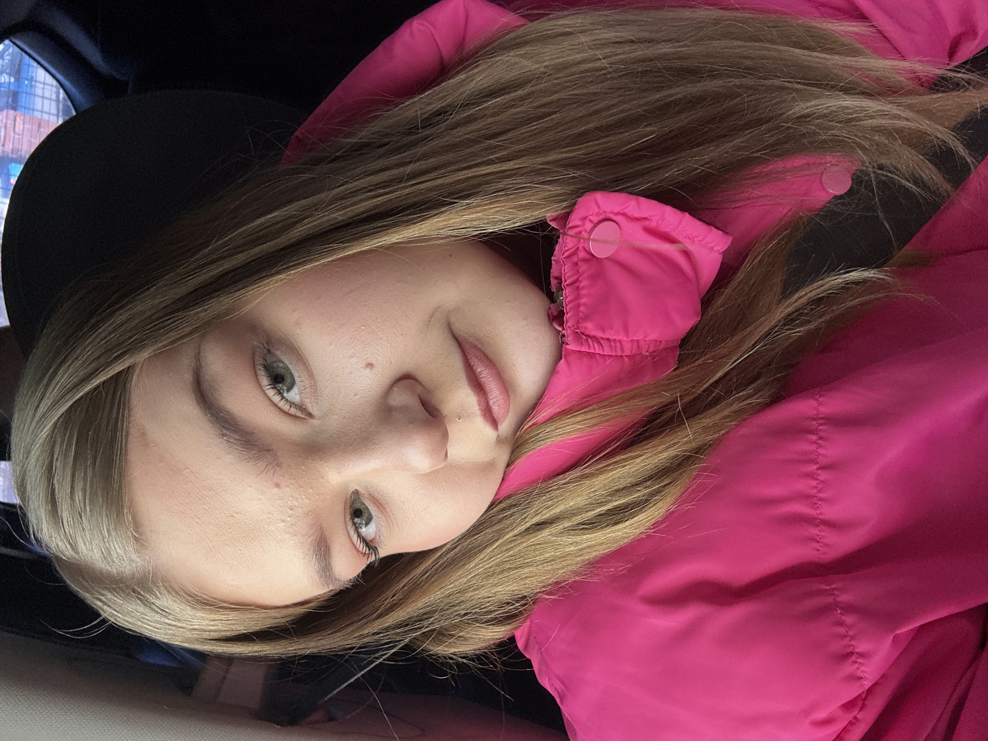 A close-up portrait features a female subject with light brown hair and green eyes, wearing a bright fuchsia puffer jacket, seated inside a vehicle in the city of Фоки, Russia. The subject's head is slightly tilted, and she looks directly at the camera with a neutral expression. Her long hair is parted on the left and falls over her shoulders and the jacket. A dark strap, consistent with a seatbelt, is partially visible diagonally across her chest beneath her hair and the jacket's collar. The background is dark and significantly out of focus, showing indistinct structural elements consistent with a vehicle interior.
