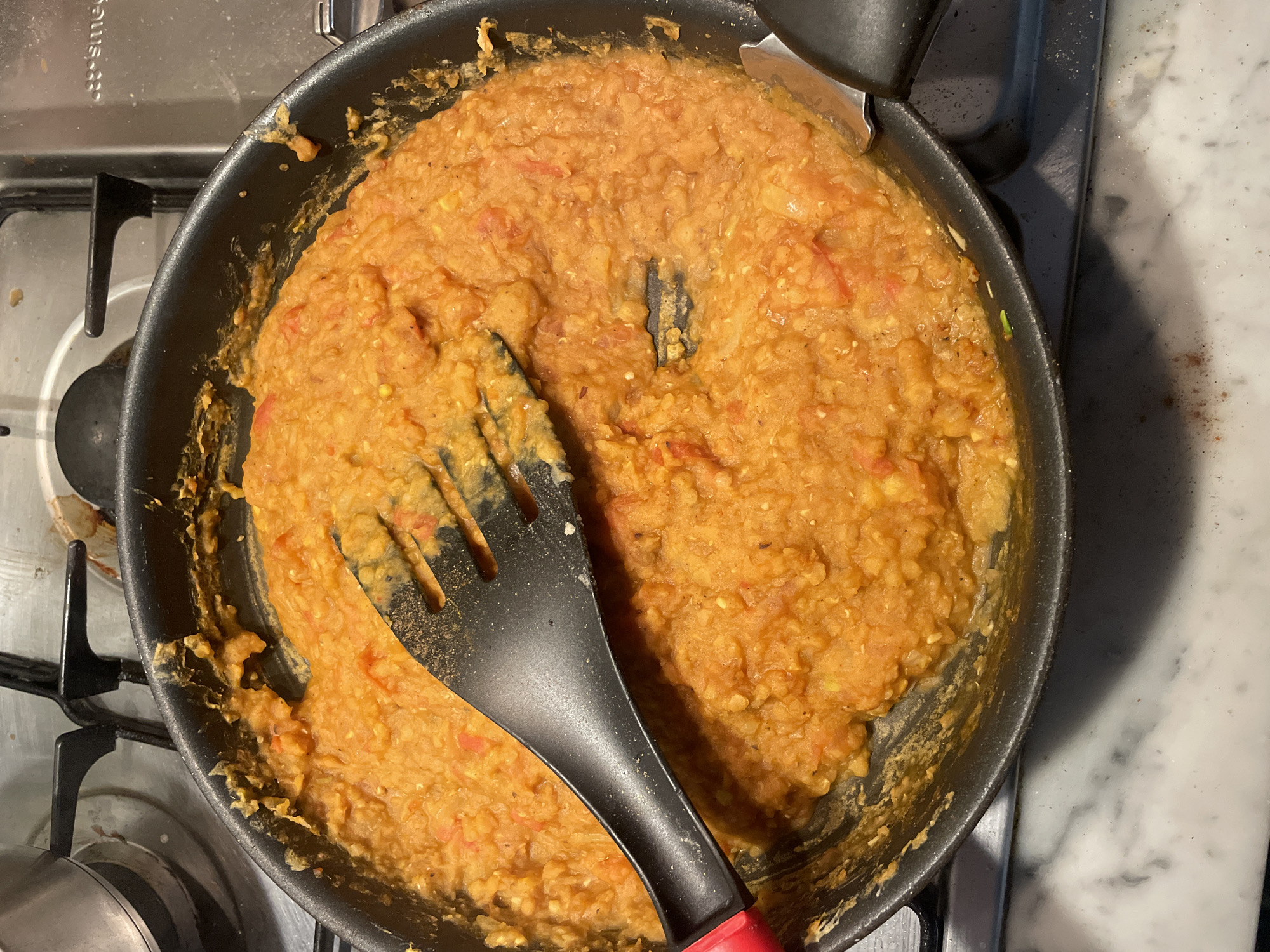 The image shows a close-up, overhead view of a black frying pan filled with a thick, orange, lentil-based stew. A black spatula with a red handle is resting on the stew, partially obscuring it. The pan is situated on a gas stovetop, with metal grates visible. The stovetop appears to be in a kitchen environment, with a stainless steel surface and some white marble countertop visible in the foreground. There are no people visible in the image. The scene is not identifiable as being in Melbourne, Australia.
