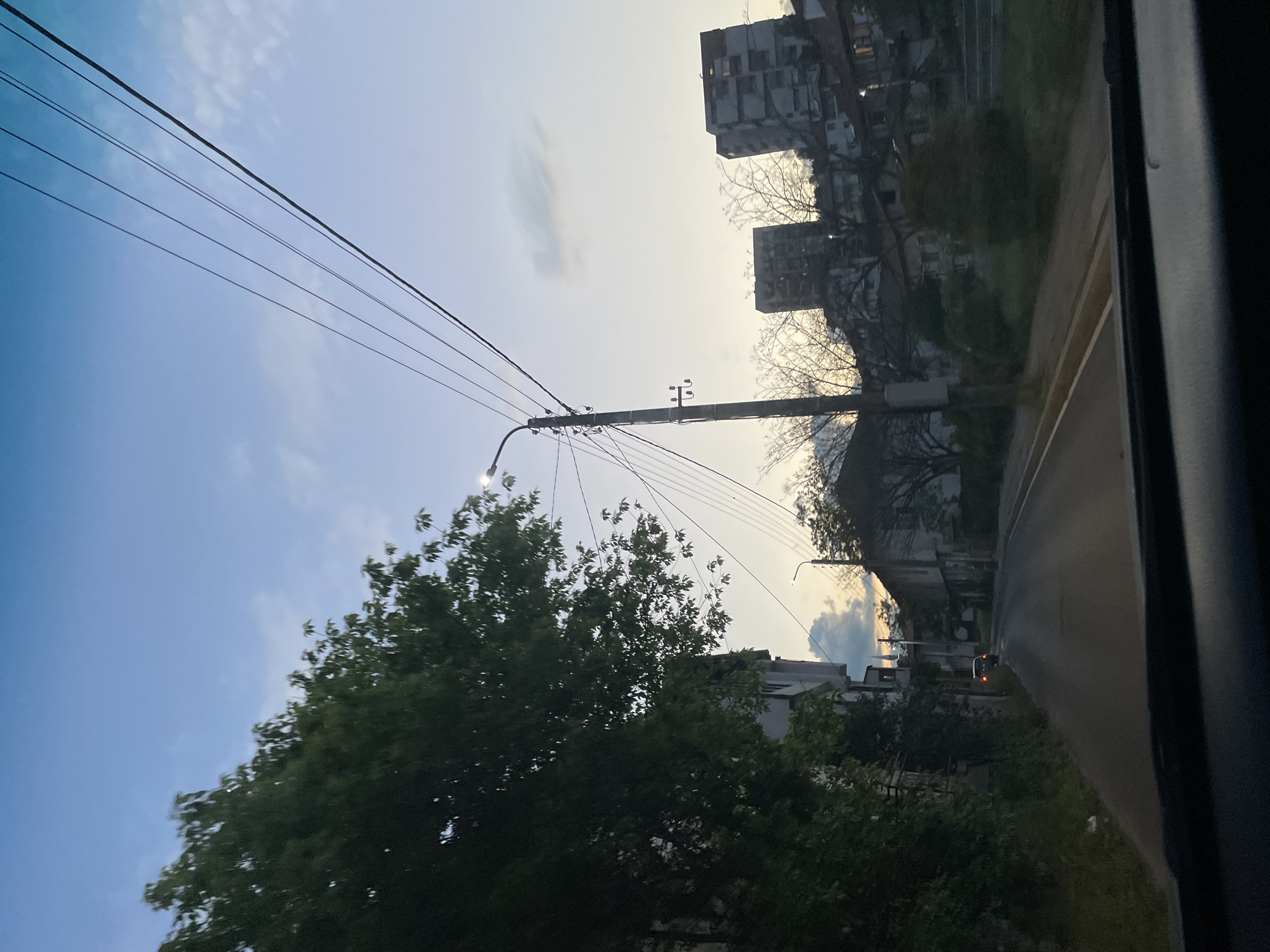 The image shows a street in Zlatograd, Bulgaria, viewed from inside a vehicle. A utility pole with wires is visible against a partly cloudy sky. Buildings line the street, obscured by trees and foliage. A road runs along the right side of the image, with a vehicle visible in the distance. The time of day appears to be dusk or dawn.