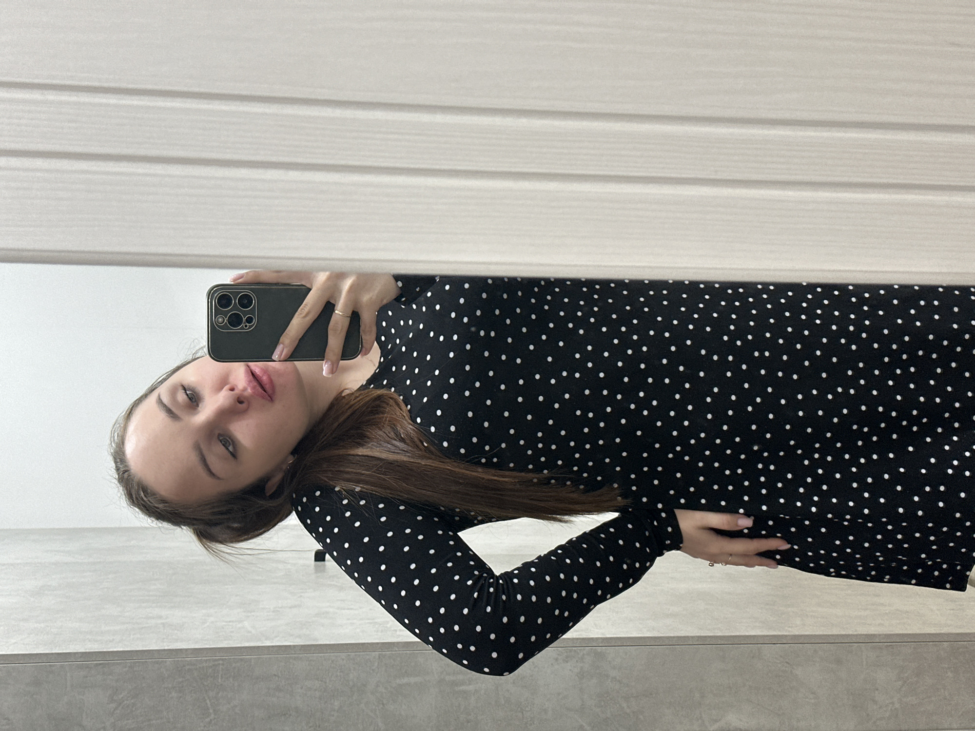 A young woman takes a selfie in a mirror. She is wearing a black dress with small white polka dots. She is holding a black smartphone with multiple camera lenses. The background includes a white slatted wall and gray tiling. The location is Chaikovsky, Russia.