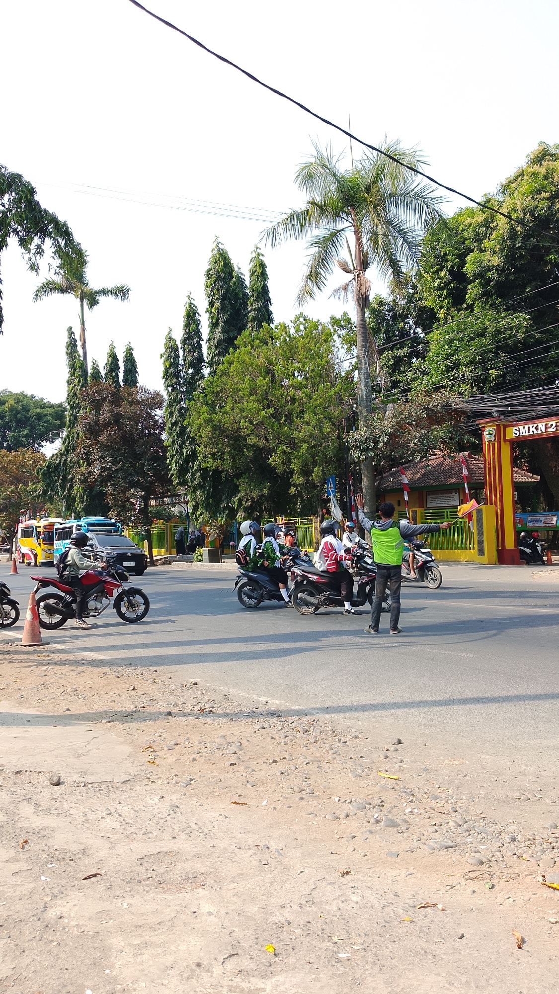 The image depicts a street scene in front of SMKN 2, a vocational high school in Procot, Indonesia. The foreground shows a dusty, unpaved roadside with scattered pebbles and dry leaves. The paved road in the middle ground is occupied by several motorcyclists, many appearing to be students wearing helmets and school uniforms. A traffic director, wearing a bright green vest, stands in the middle of the road, gesturing with their hands, possibly directing traffic or guiding students. In the background, a yellow and red school gate with the inscription "SMKN 2" is visible, along with trees and a cloudy sky. Several colorful buses are also present on the road, suggesting school transportation. The overall impression is a busy school entrance during daytime.