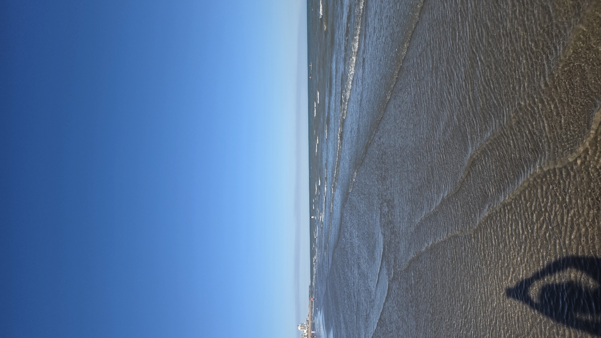 A wide shot of a calm beach under a clear blue sky. The foreground features shallow water with visible ripples from gentle waves lapping at the shore. A shadow of a person in a yoga pose is cast on the wet sand in the lower left corner. In the mid-ground, the ocean stretches to the horizon with small waves. In the distance, along the shoreline, a few people are visible walking and standing, and a pier extends into the sea from the left. A few buildings are also discernible on the coastline to the far left. The scene appears to be St Leonards, United Kingdom.