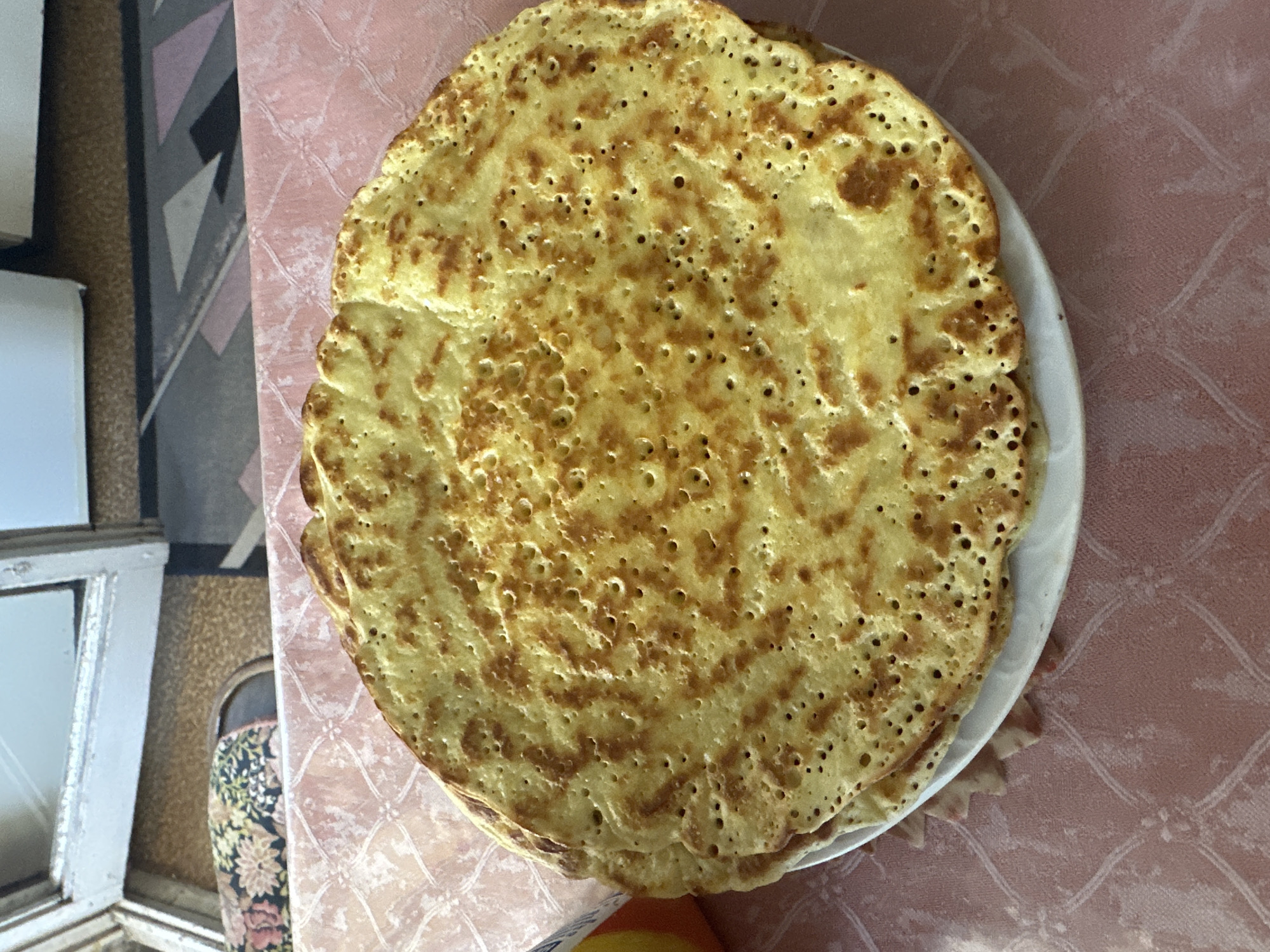 The image displays a stack of approximately 5-7 thin, golden-brown pancakes or crepes, exhibiting an irregular porous surface pattern from cooking, centered on a white, round plate. The plate rests on a pink tablecloth with a subtle, embossed repeating pattern. In the upper-left background, a worn, white-painted wooden window frame is visible, beyond which a patterned slipper or soft shoe with a dark floral design on a light brown floor is partially seen. The upper-right background reveals interior elements, including a white object (possibly furniture or an appliance) and a geometric patterned floor covering in shades of grey and black. A small, partially visible yellow and white object is situated to the left of the plate. The scene takes place in the city of Svilengrad, Bulgaria. No individuals or specific actions are depicted.