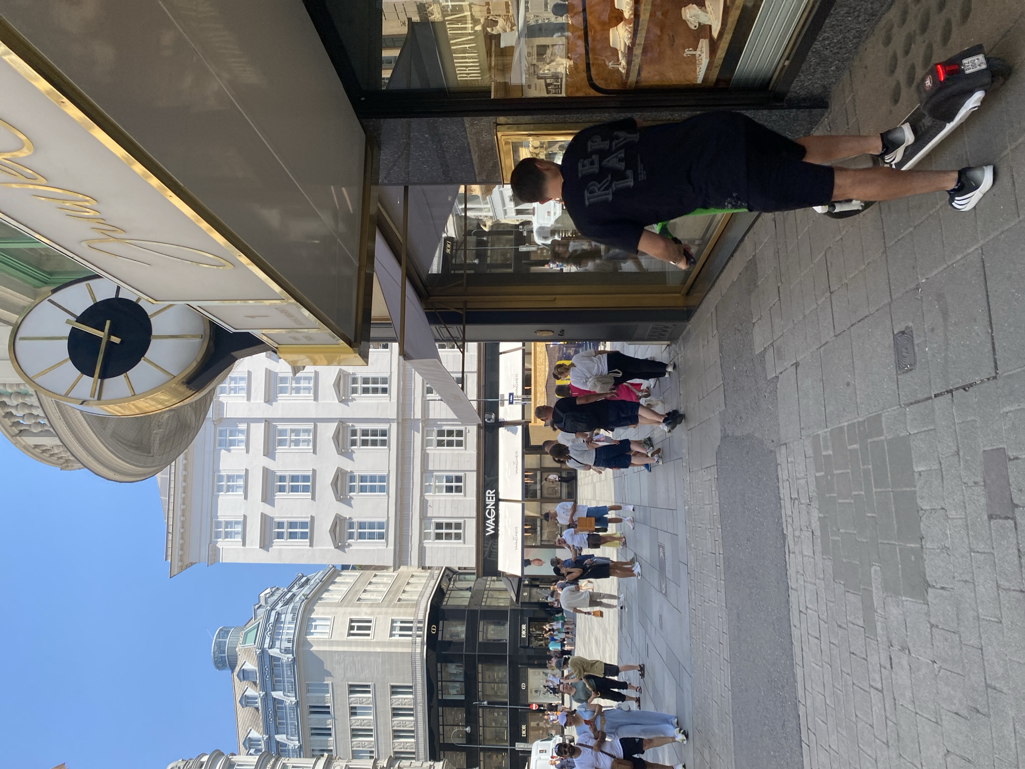 The image depicts a street scene in Vienna, Austria, likely during daytime hours given the bright sunlight. In the foreground, a person is riding a lime green electric scooter on a cobblestone sidewalk. The rider is wearing a black t-shirt with the word "REPLAY" printed on the back in a distressed font, black shorts, and black and white Adidas sneakers.
The background reveals a bustling city environment with pedestrians walking on the sidewalk. Several shops are visible, including a "DIOR" store and a shop with the name "WAGNER" above its entrance. Another store has a sign that reads "JUWELEN" (meaning jewelry in German) in a golden script, accompanied by a large clock. Buildings in the background are tall and architecturally diverse, some appearing to be historic. The sky is clear and blue. The scene captures a moment of urban life, with people shopping, walking, and commuting.