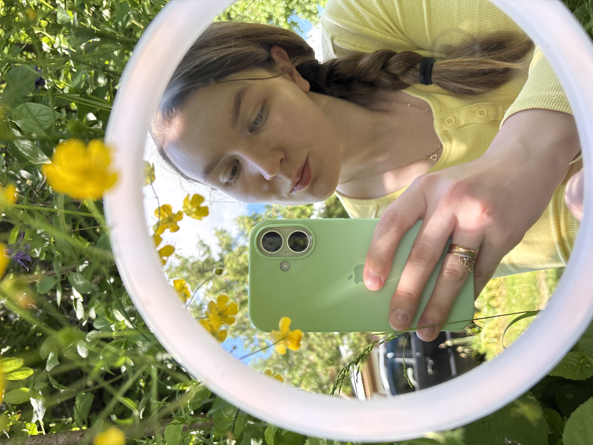 The image presents a close-up, outdoor scene captured in Italmas, Russia, centered on a circular mirror with a thick, white frame. The mirror reflects a young woman in the act of taking a self-portrait with a smartphone.
The woman has light skin, brown hair styled in a side braid secured with a black elastic band, and is wearing a light yellow, ribbed, long-sleeved Henley-style shirt with visible yellow buttons. A delicate gold chain necklace with a small heart-shaped pendant is worn around her neck. Her right hand, visible on the left side of the reflection, holds a light green smartphone, identifiable by its dual-camera setup and an Apple logo on the case. Two gold rings are visible on her right ring finger: a plain band and an ornate, possibly crown-shaped, ring. Her gaze is directed downwards towards the phone.
The environment outside the mirror is characterized by lush green vegetation, including various leaves, plants, grasses, and a tree trunk visible on the left. Scattered throughout the greenery are numerous bright yellow flowers, possibly buttercups, and one distinct purplish-blue flower in the upper left background. The scene is brightly illuminated, suggesting a sunny day.
Within the mirror's reflection, behind the woman, an open, bright blue sky with some white clouds is visible above a backdrop of green trees and foliage. More yellow flowers are also reflected in the background. In the lower-left quadrant of the reflection, a dark-colored vehicle, possibly a car or SUV, is partially visible, indicating a nearby road or parked area within the otherwise natural setting.